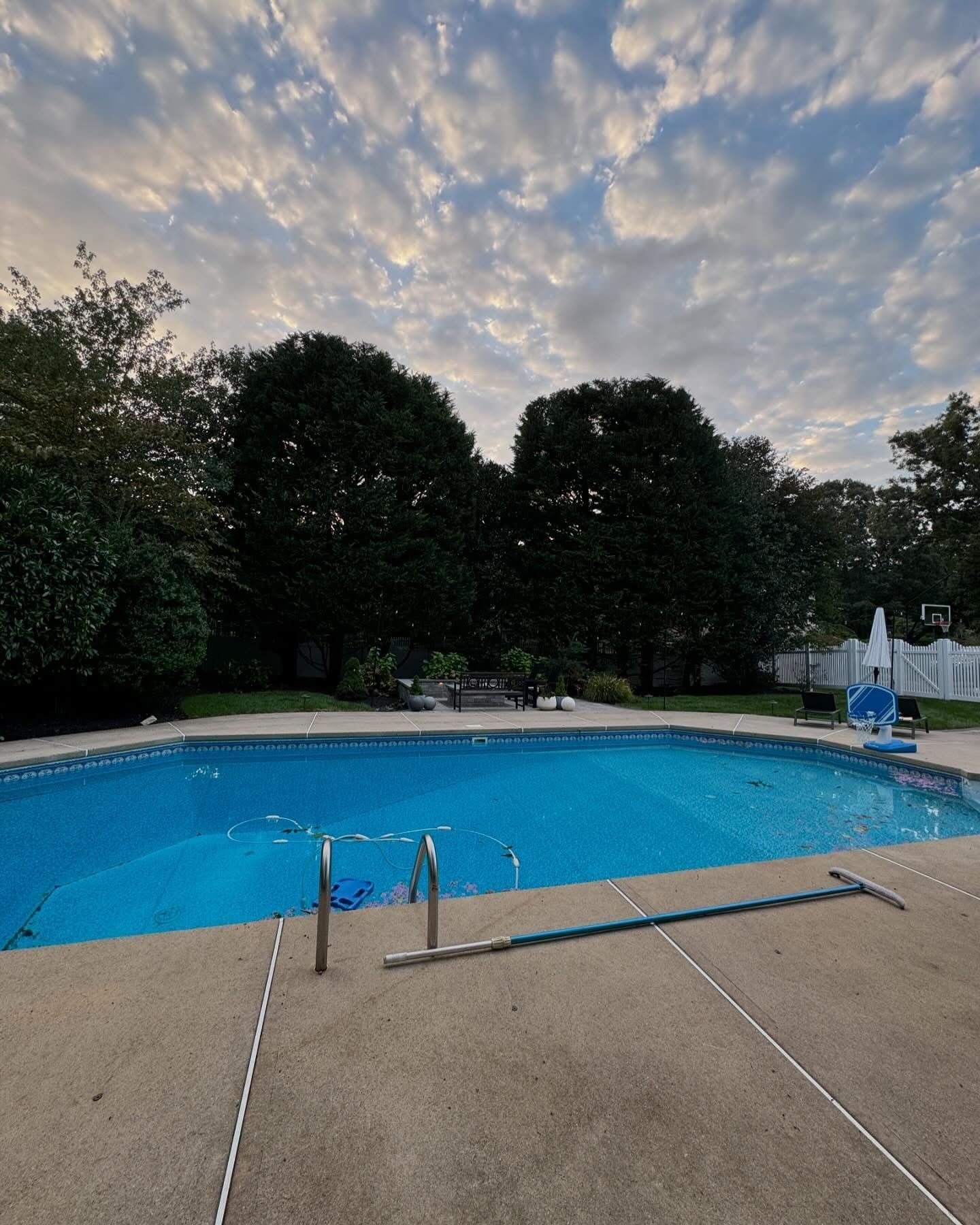 Empty outdoor swimming pool with blue water, surrounded by trees under a cloudy sky at dusk