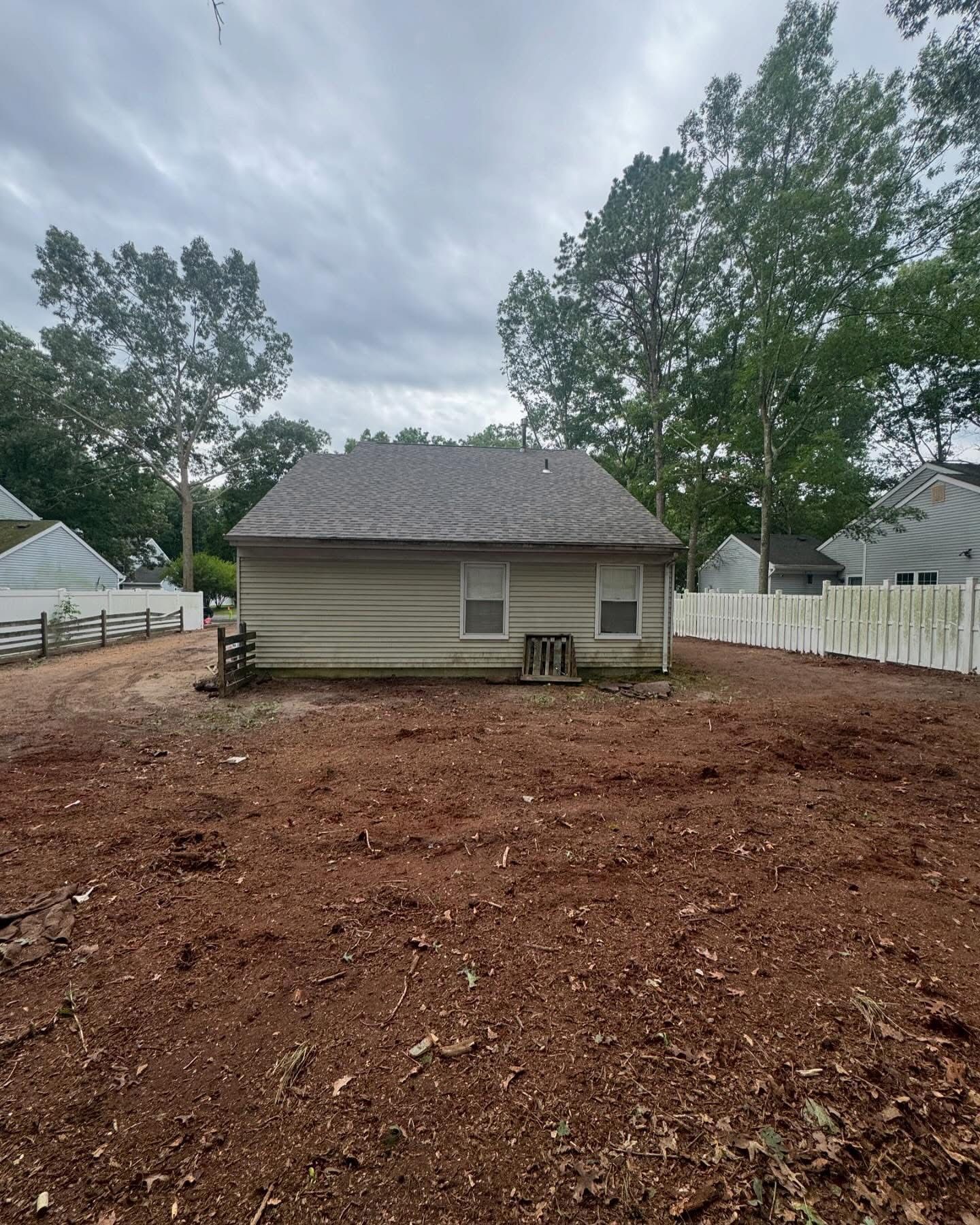 Small beige house with a gray roof beside a white fence, surrounded by a bare dirt yard under cloudy skies.