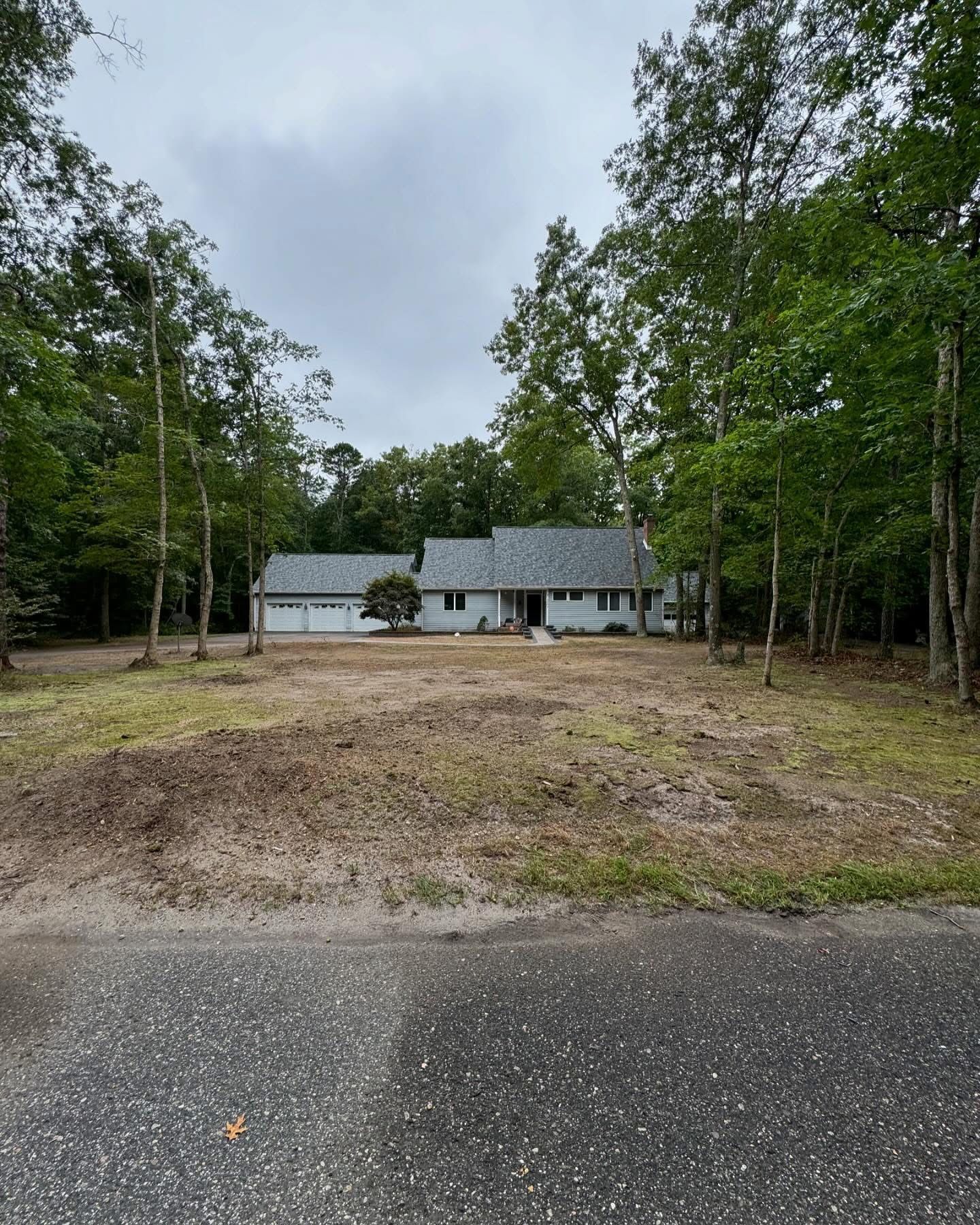 Single-story gray house in a wooded clearing with a gravel driveway and unfinished front yard.
