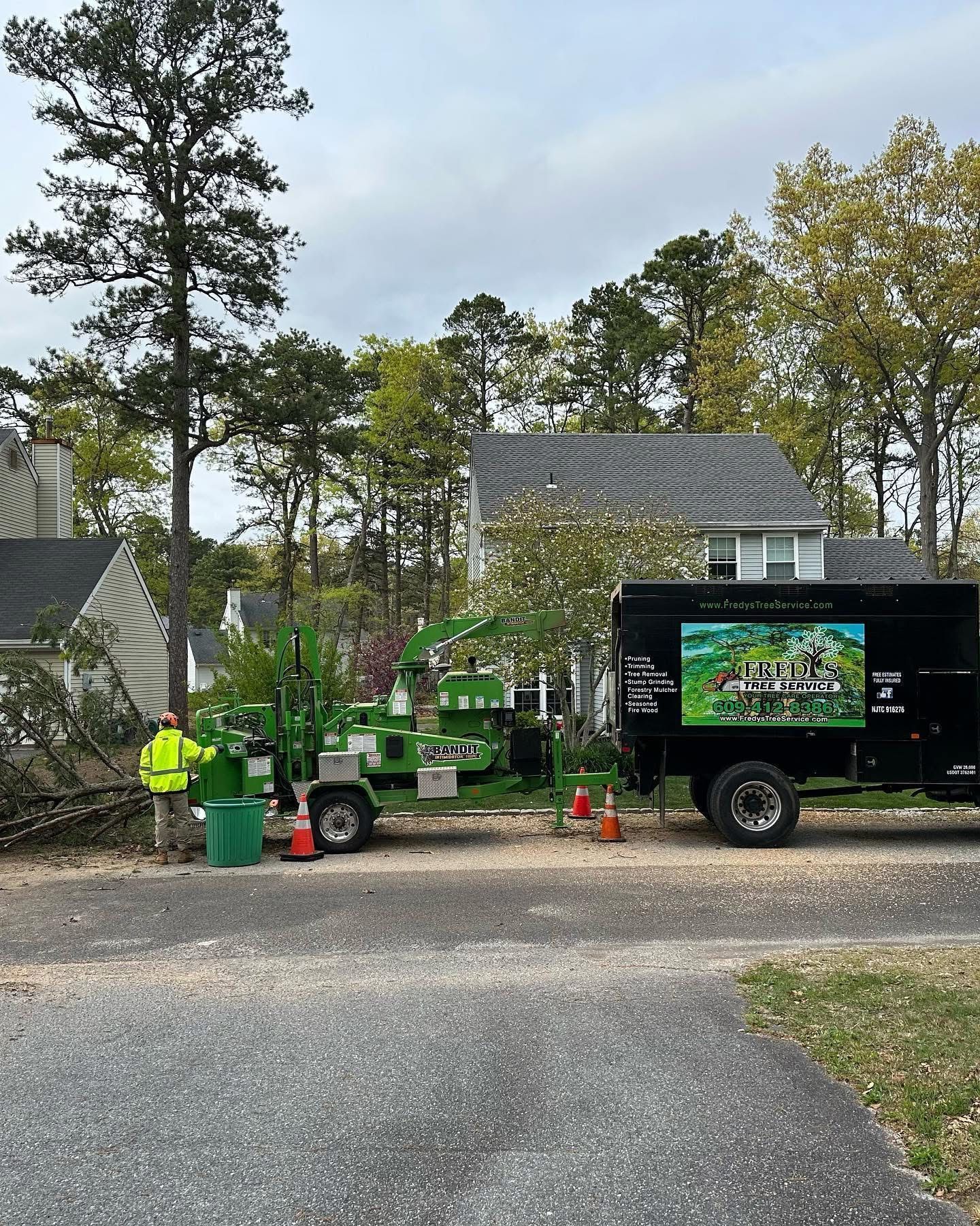 Workers with green machinery clearing debris beside a black truck in a wooded neighborhood driveway