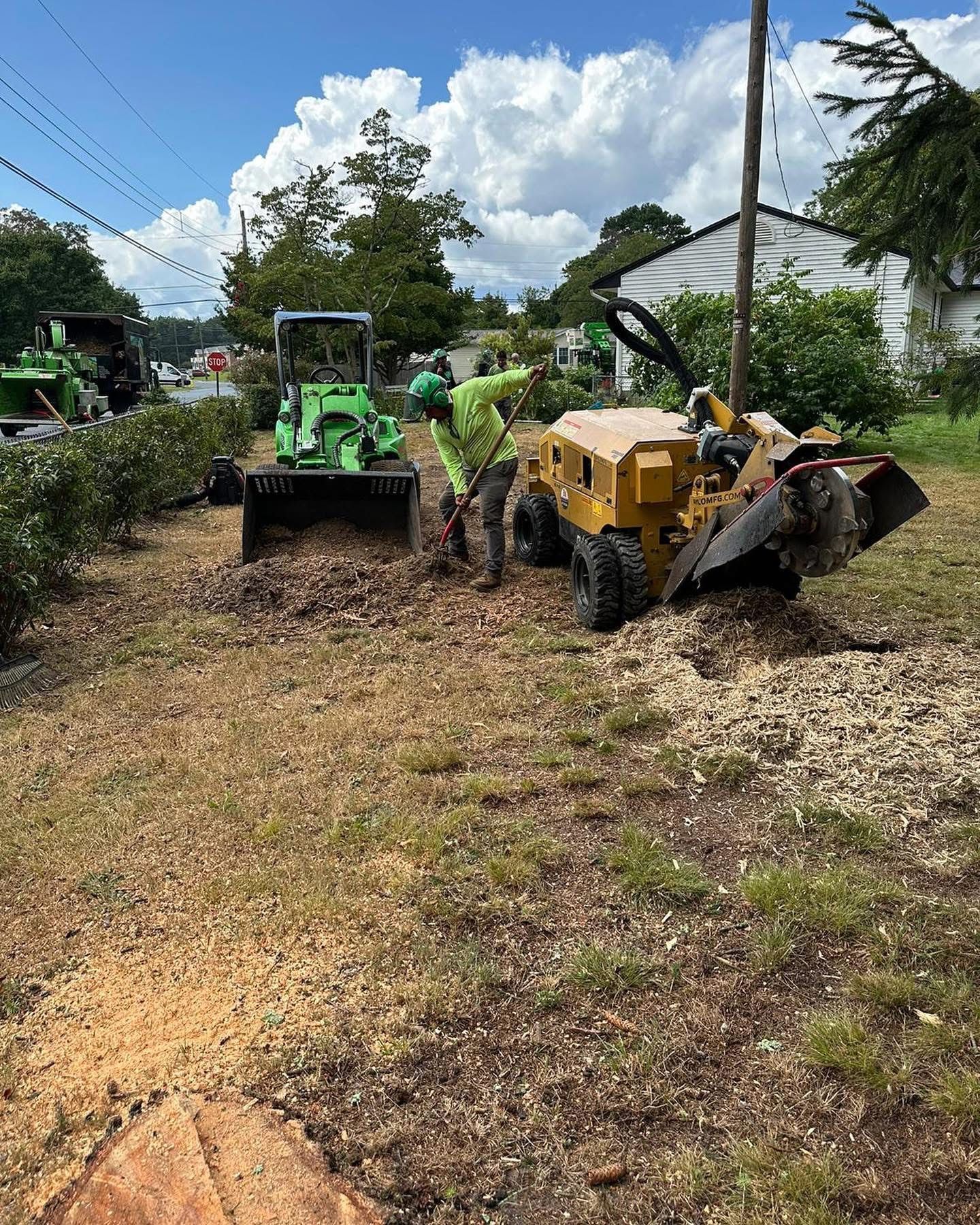 Workers using a green tractor and yellow wood chipper on a roadside, clearing brush under blue sky