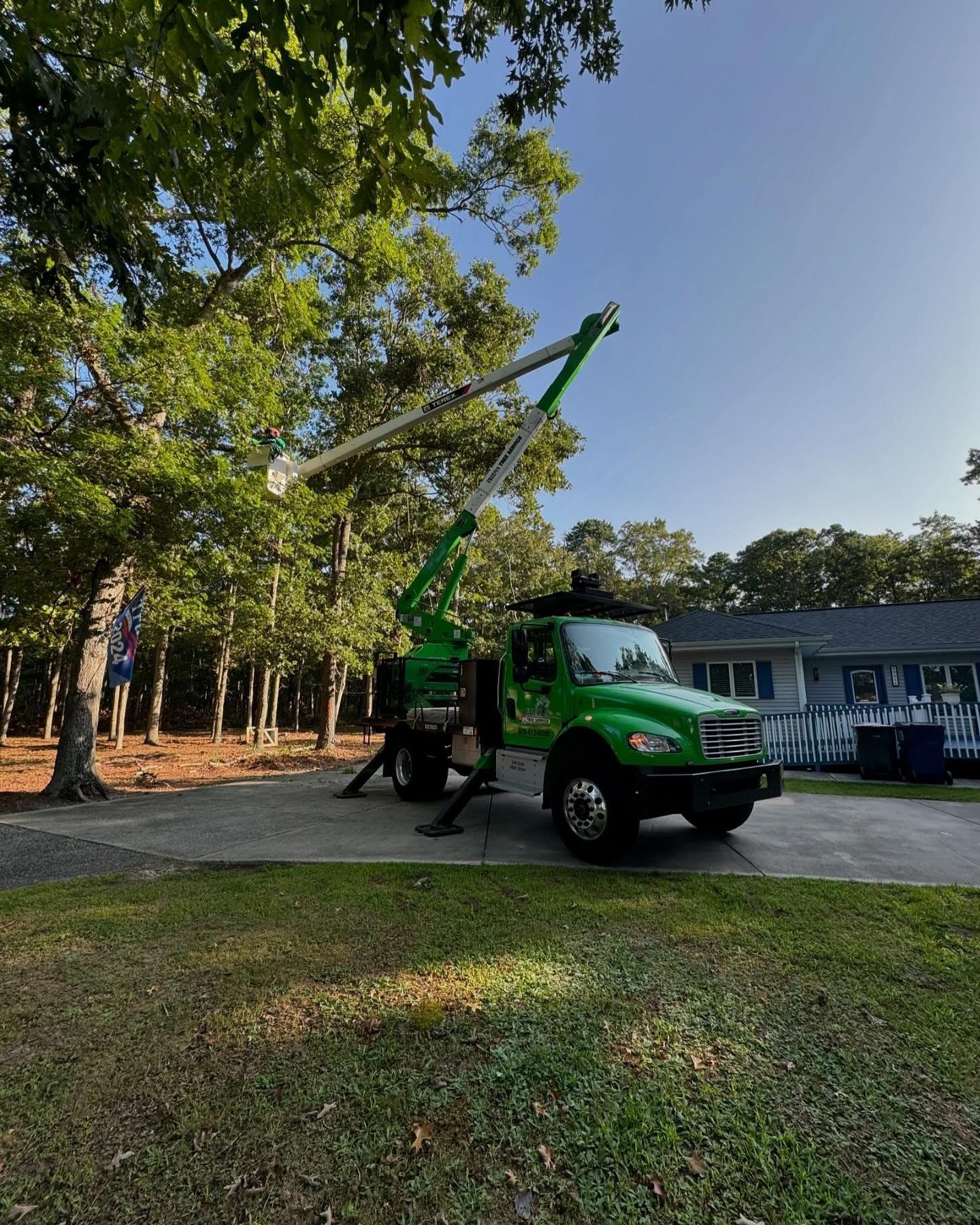 Green utility truck with a raised boom parked on a tree-lined road in daylight