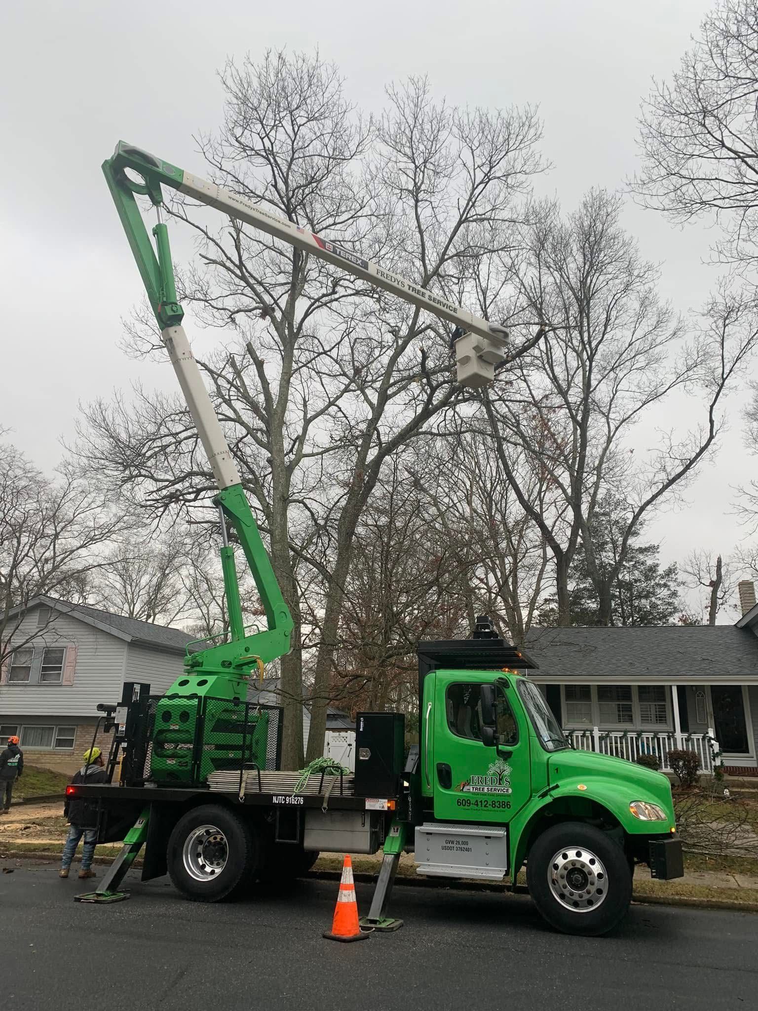 Green utility truck with raised boom parked by a house, with orange cones in a residential street