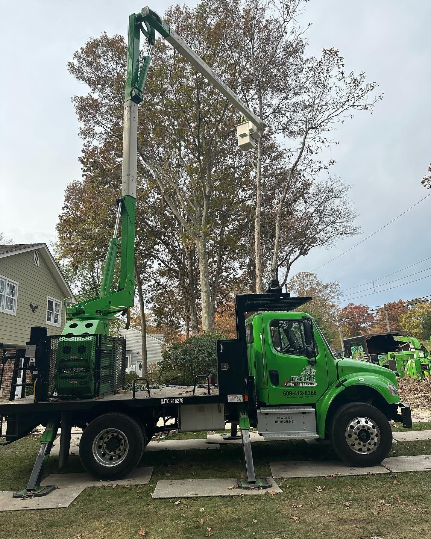 Green tree service truck with bucket lift parked on a street beside houses and autumn trees