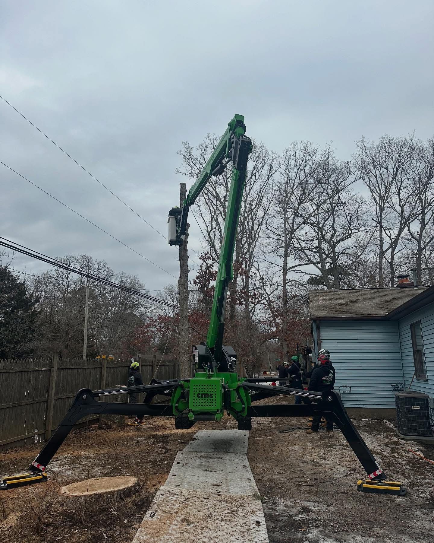 Green spider lift parked on a driveway beside a house, with trees and cloudy sky in the background.