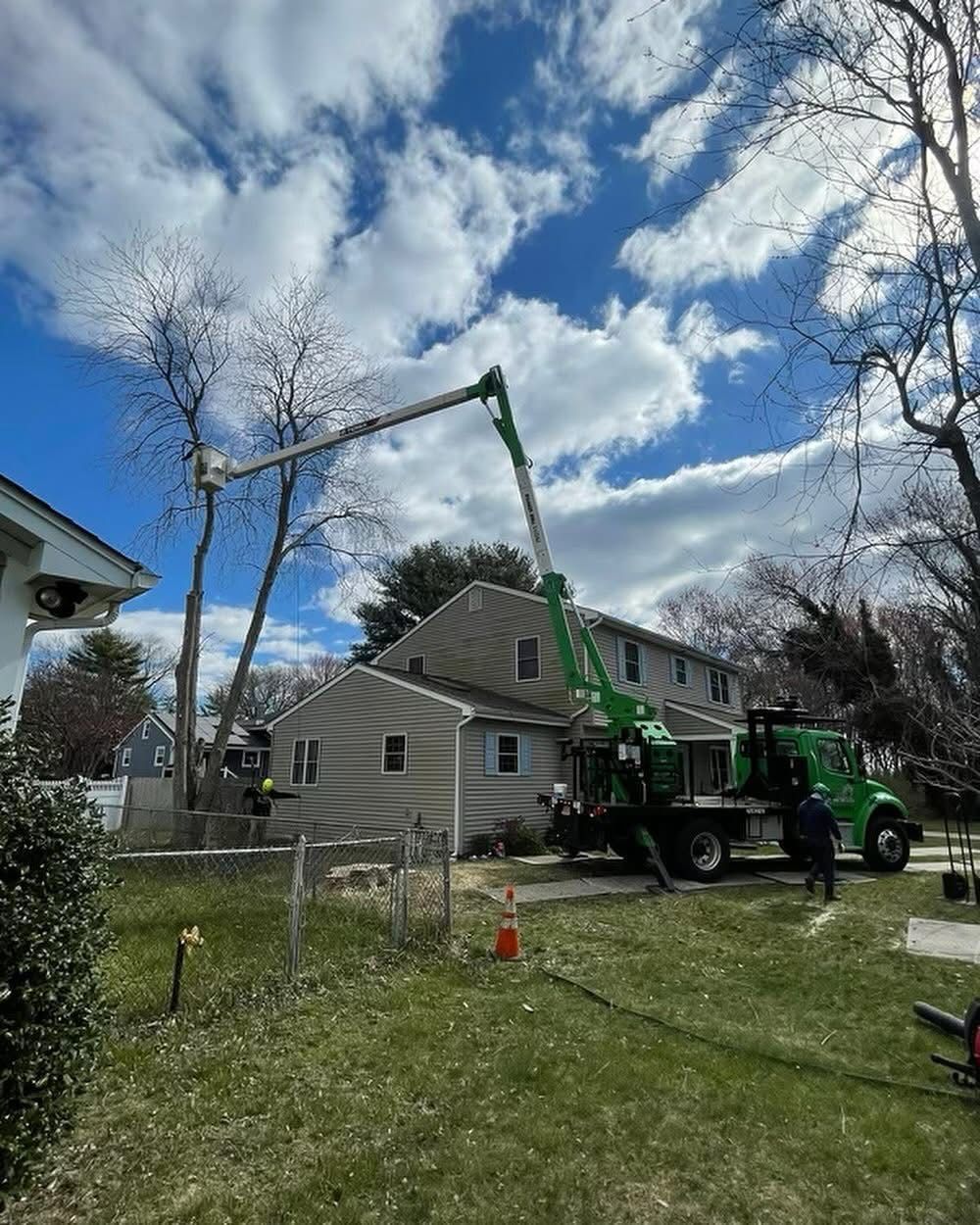 A green crane truck lifts a tree branch beside a house under a cloudy sky.