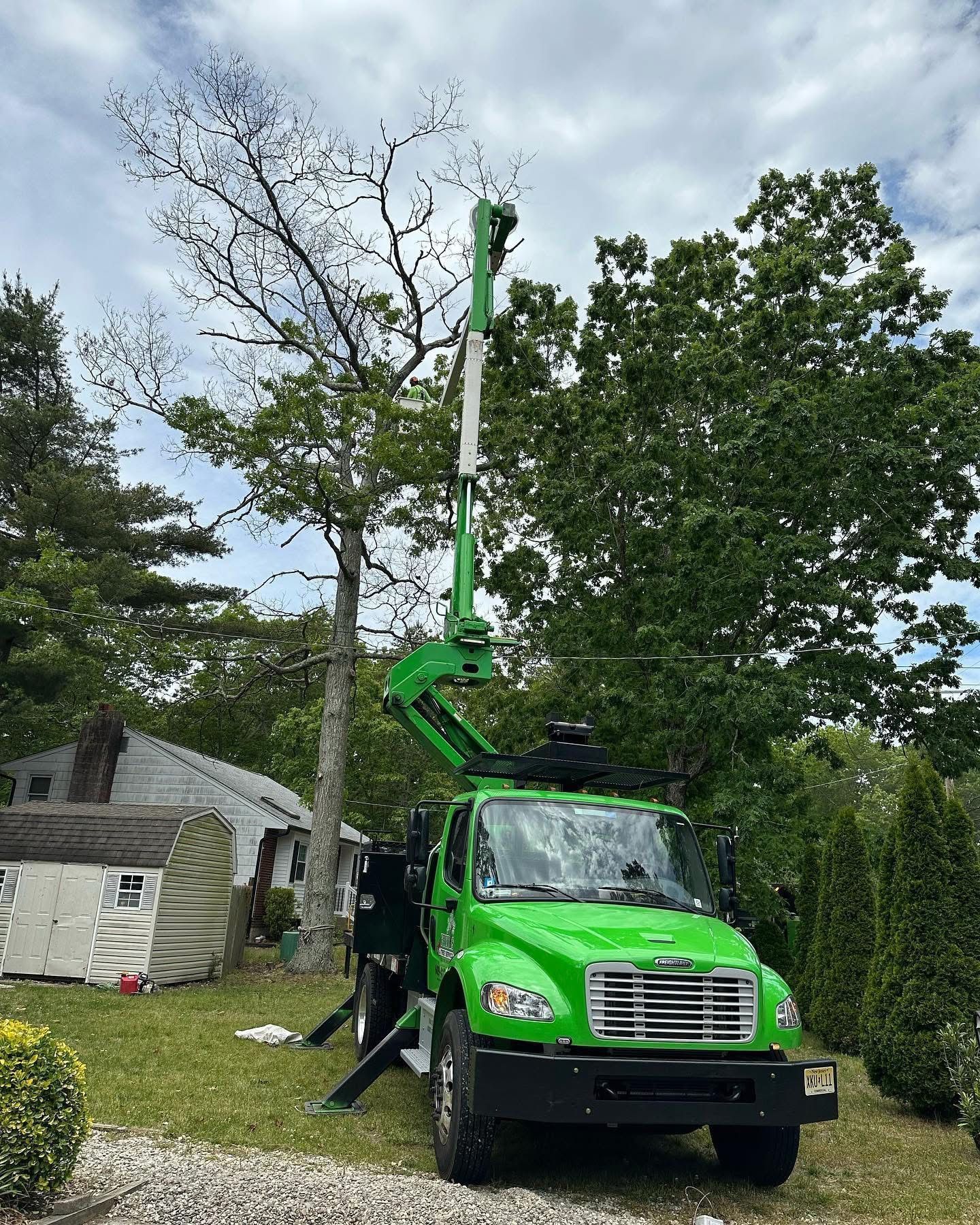 Green utility truck with raised boom parked in a yard beside a house and trees.