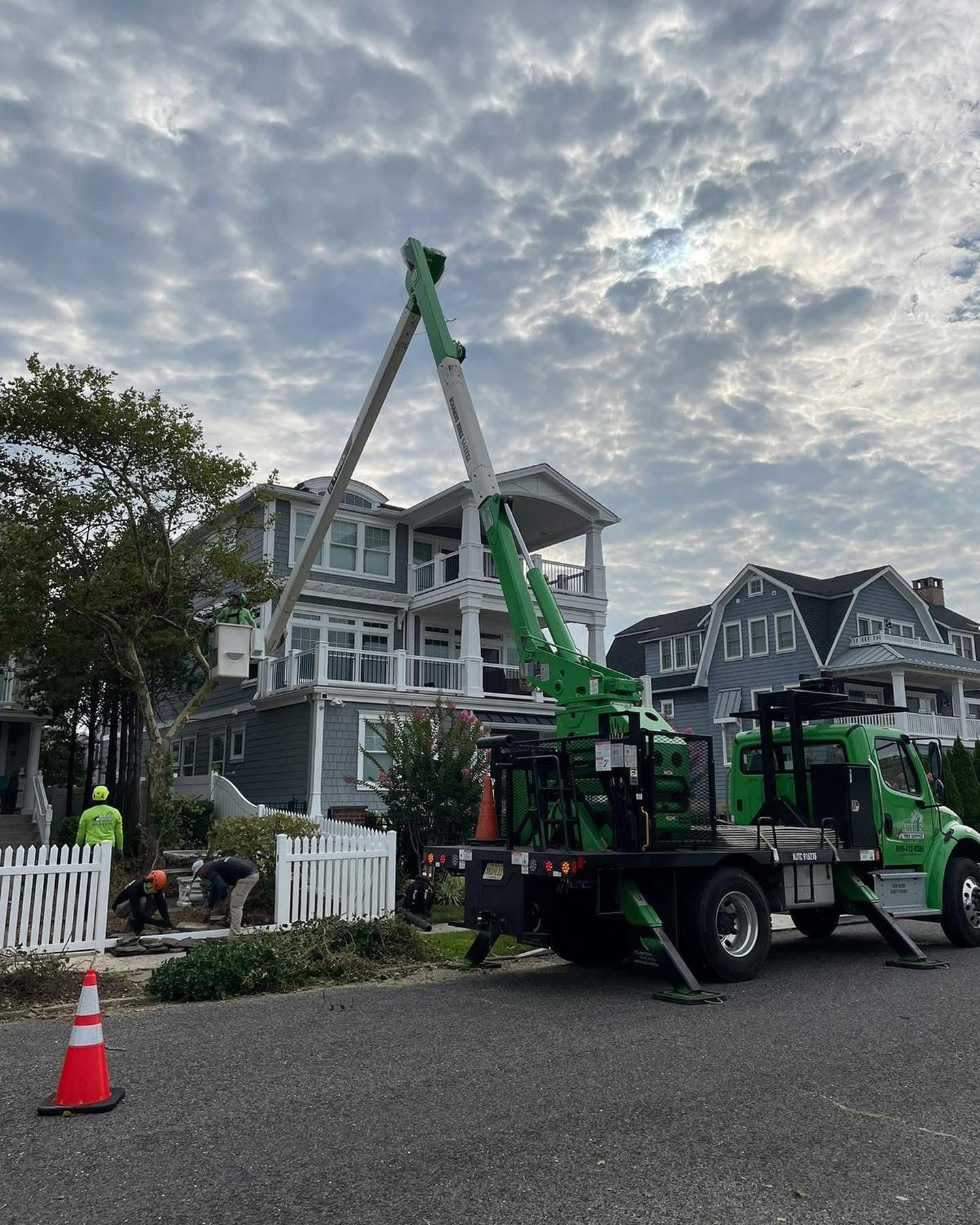 Green crane truck lifting a large tree beside a gray house on a cloudy residential street