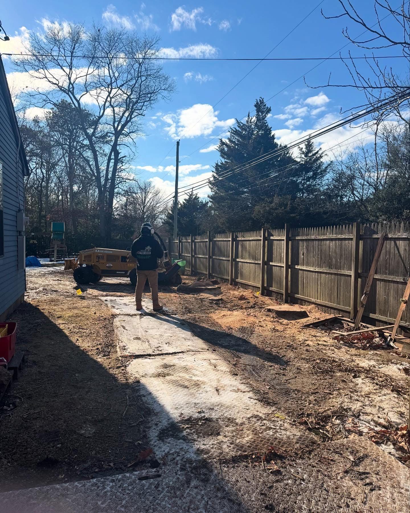 Person and dog on a sunny backyard path beside a wooden fence and leafless trees.