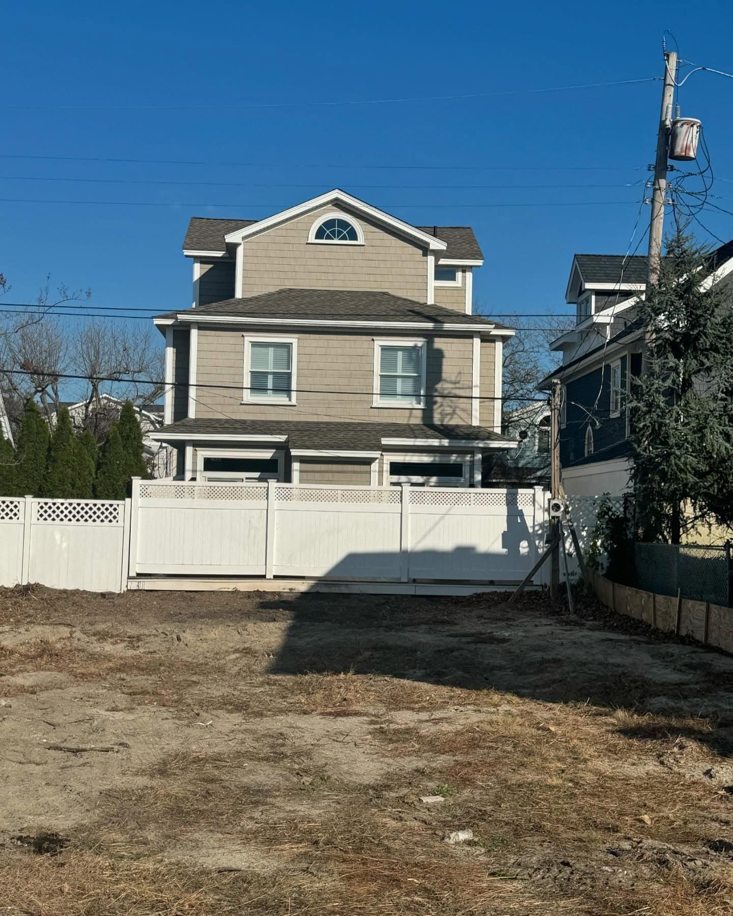 Two-story beige house with white fence in a bare yard under a clear blue sky