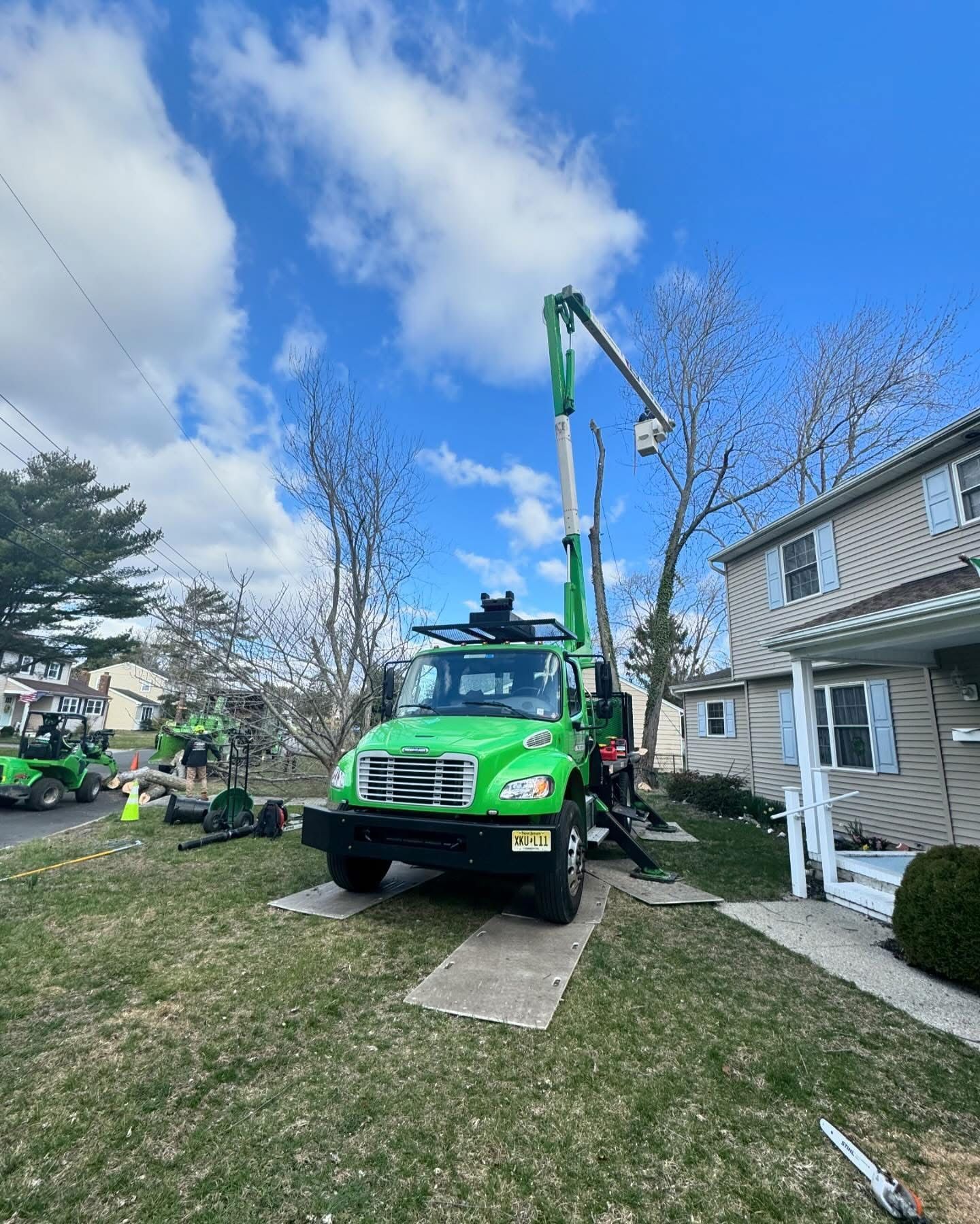Green crane truck parked beside a house, lifting near the roof on a sunny day.