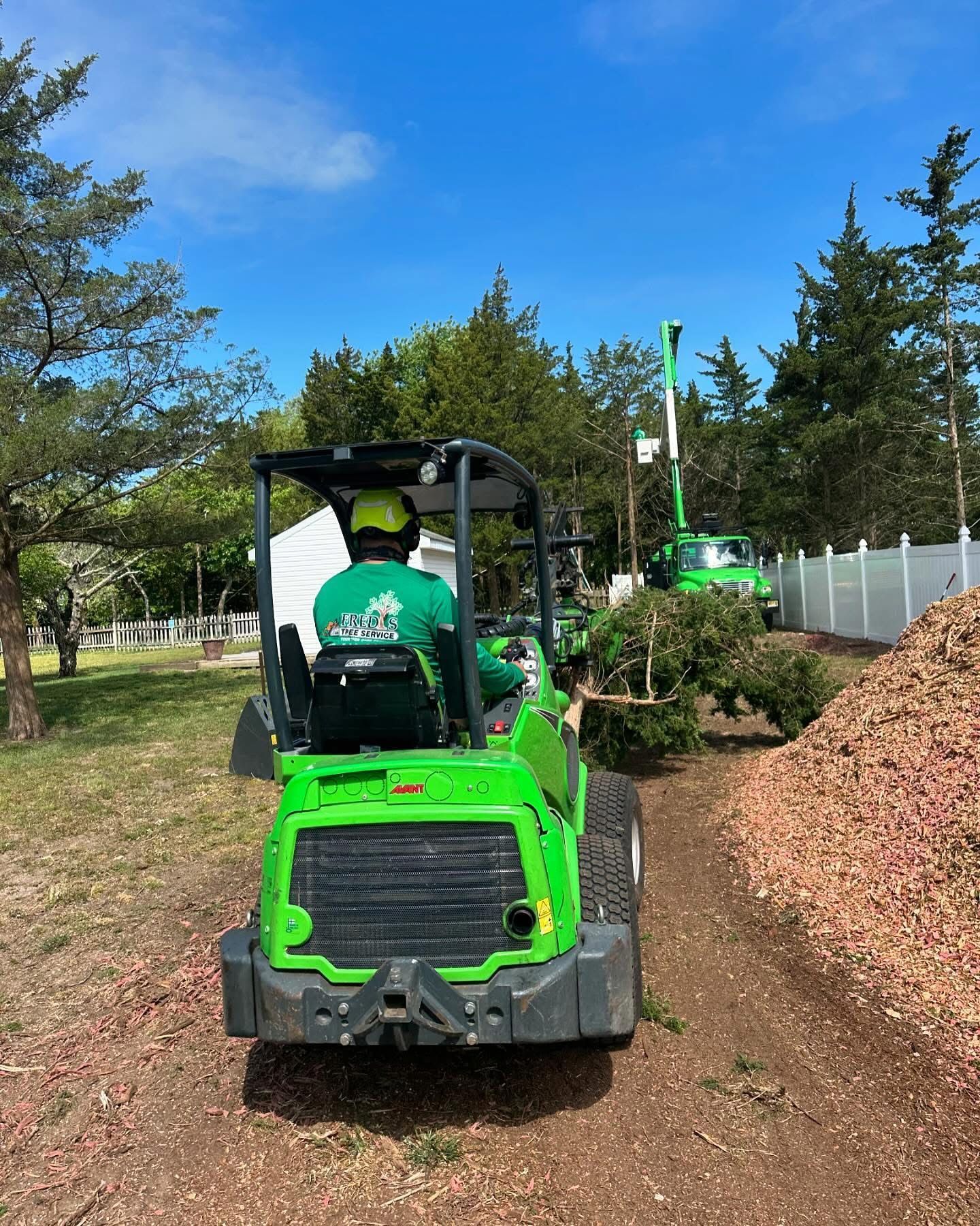 Green utility vehicle driving on a dirt path in a park, with trees, a fence, and piles of leaves nearby.