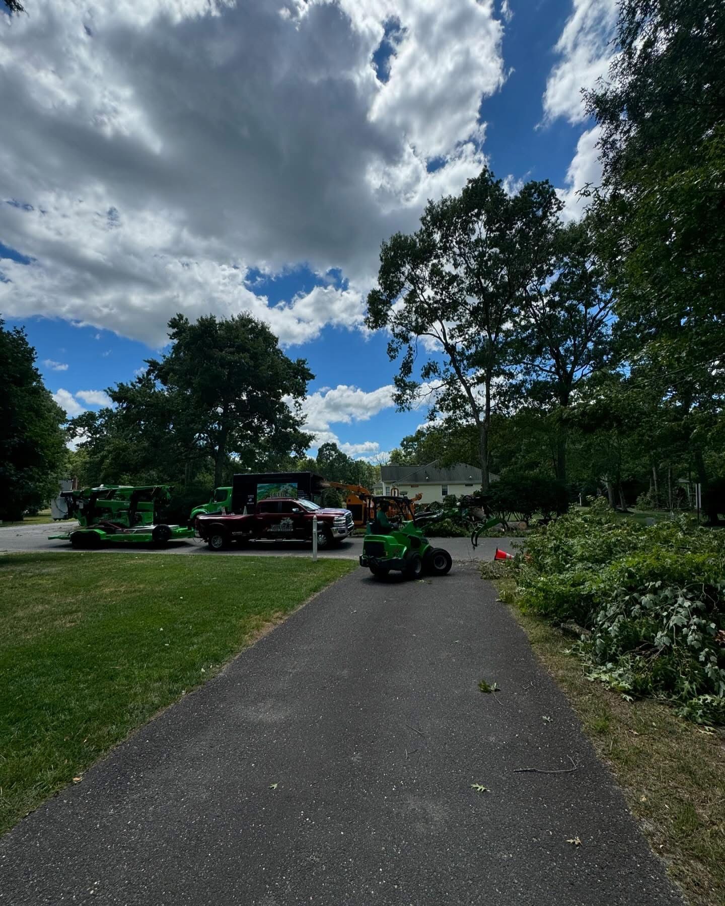Person on a green mower on a paved park path under a cloudy sky, with trees and grass around.
