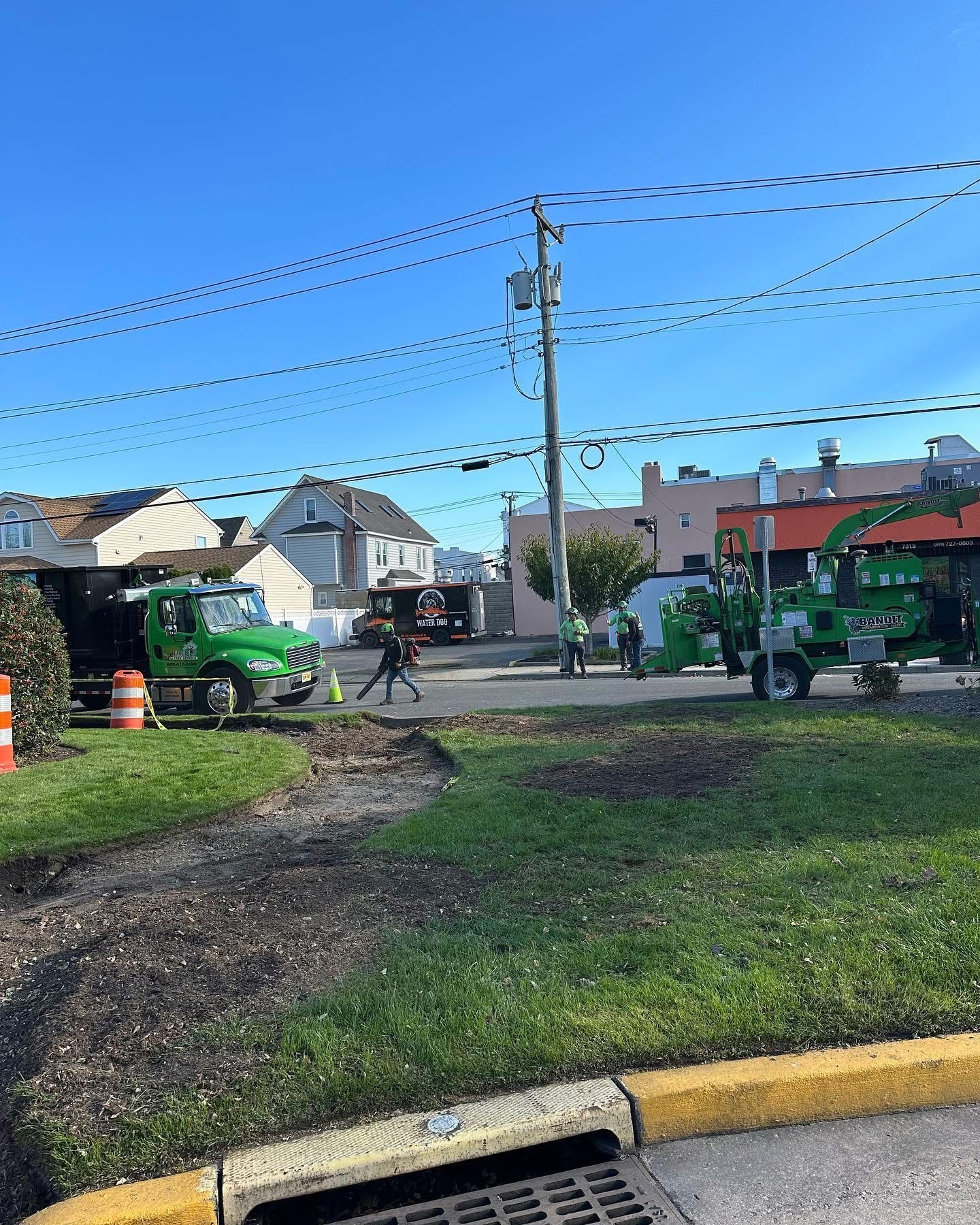 Street corner with a green utility truck, pedestrians, and houses under a clear blue sky.