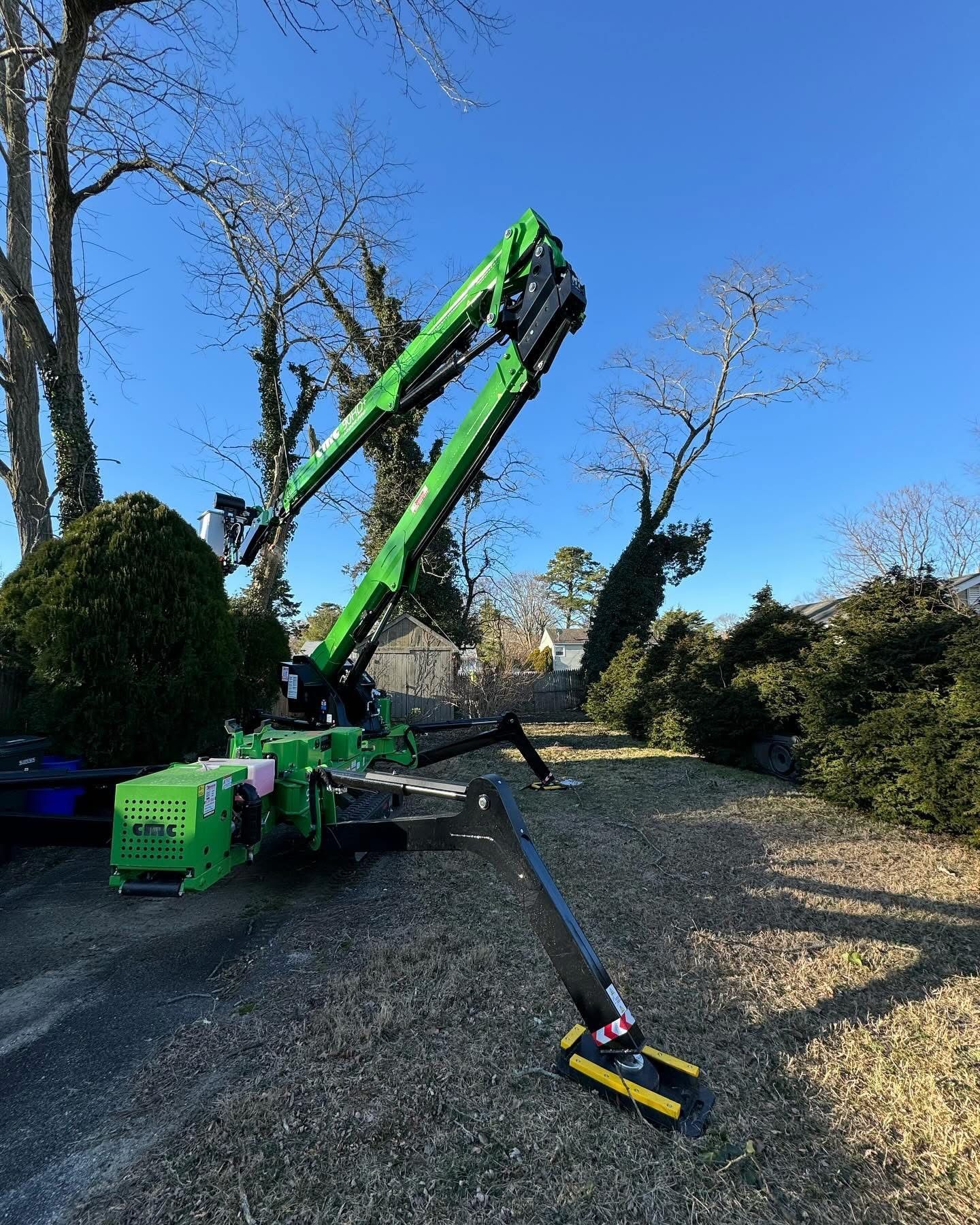 Green excavator on gravel driveway using an extended boom to trim or clear branches under blue sky.