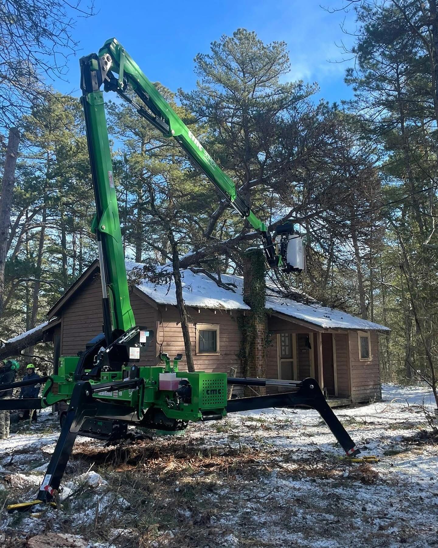 Green crane lifting a tree beside a snow-dusted log cabin in a wooded yard