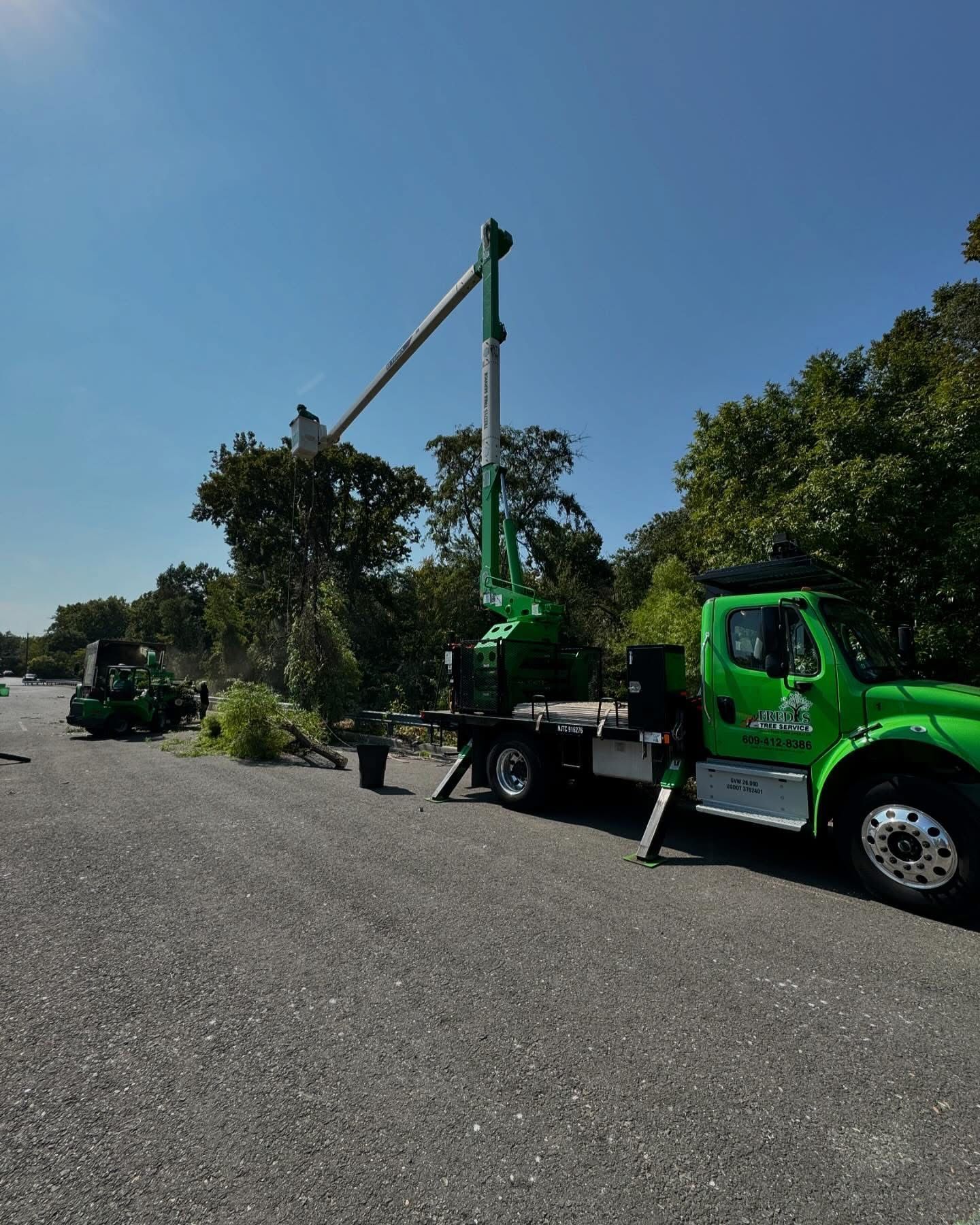 Green utility truck with extended boom parked on a sunny roadside with trees and open sky