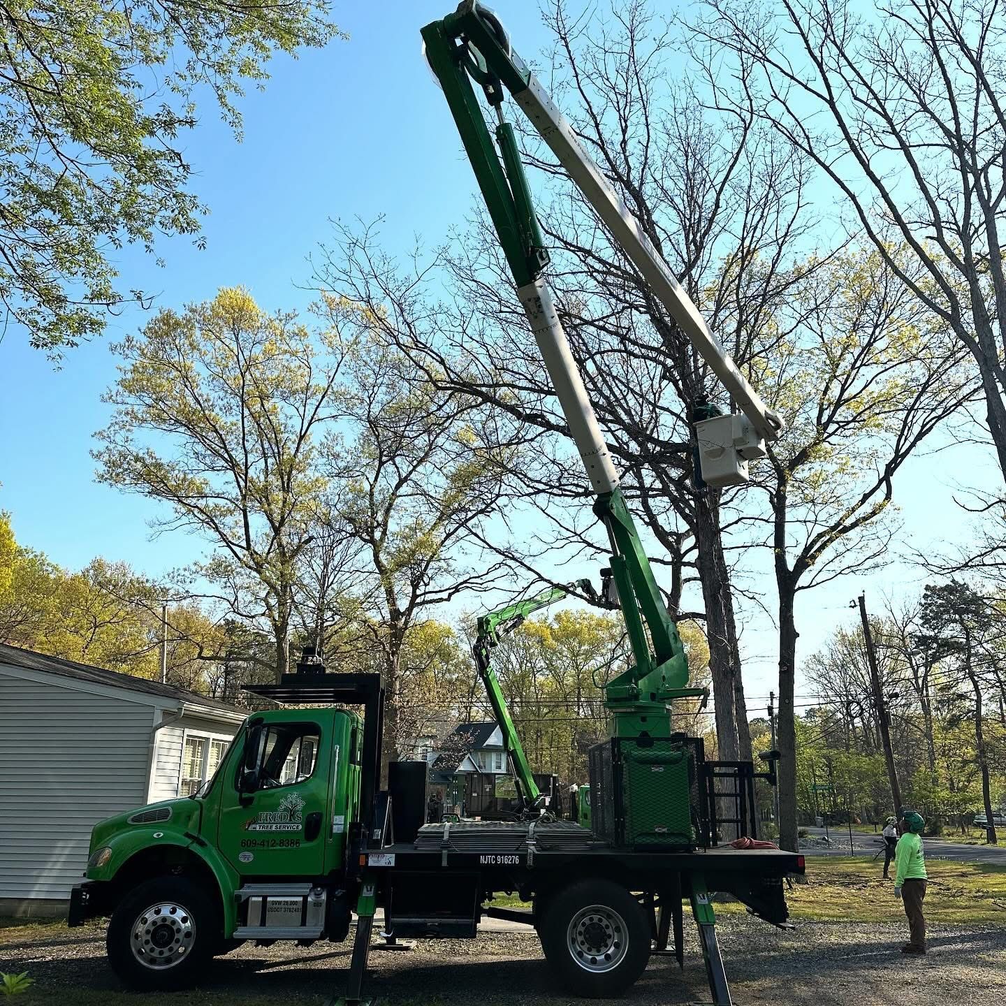 Green utility truck with extended boom lift parked on a tree-lined street, workers nearby