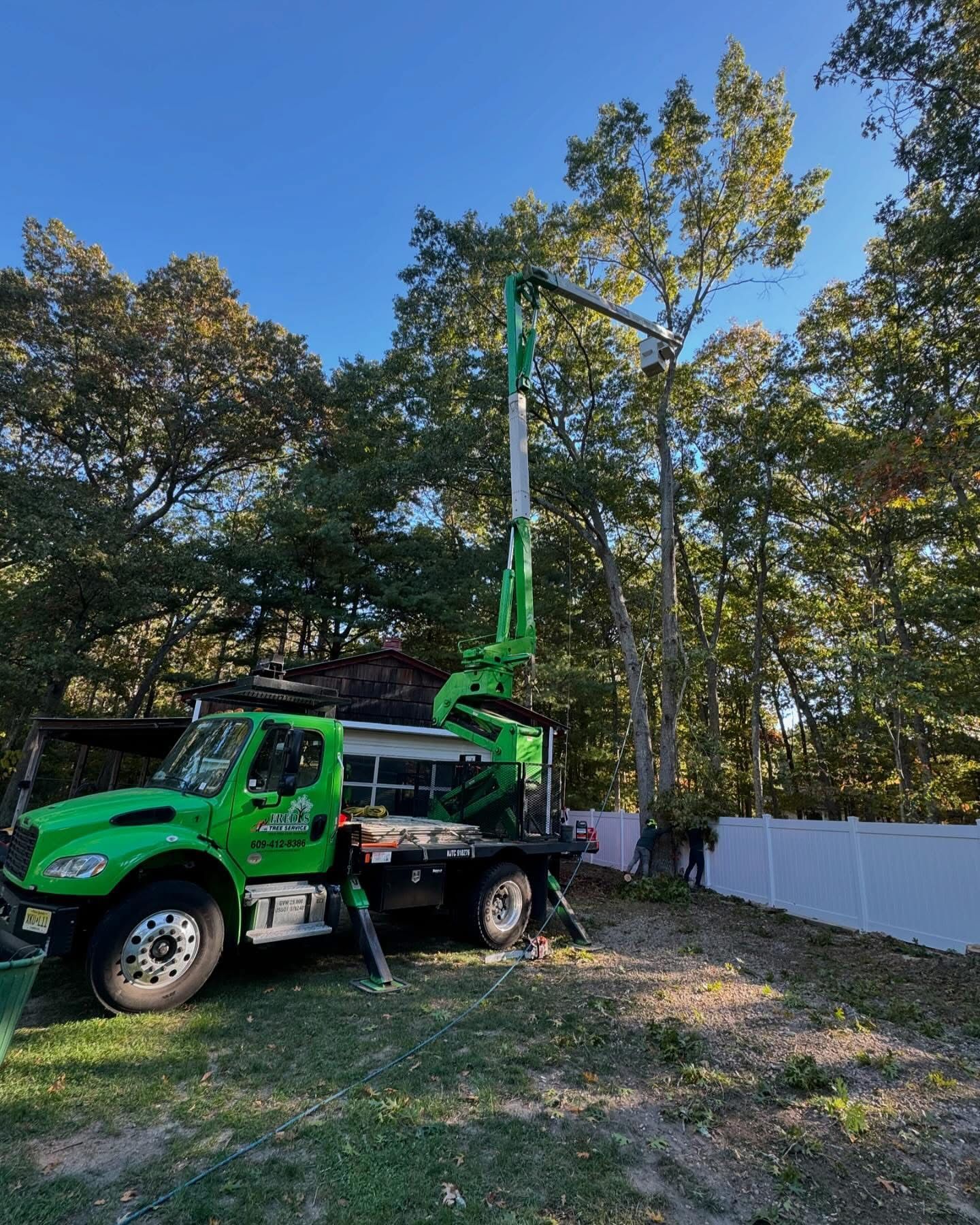 Green utility truck with raised boom parked in a grassy yard beside a white fence and trees.