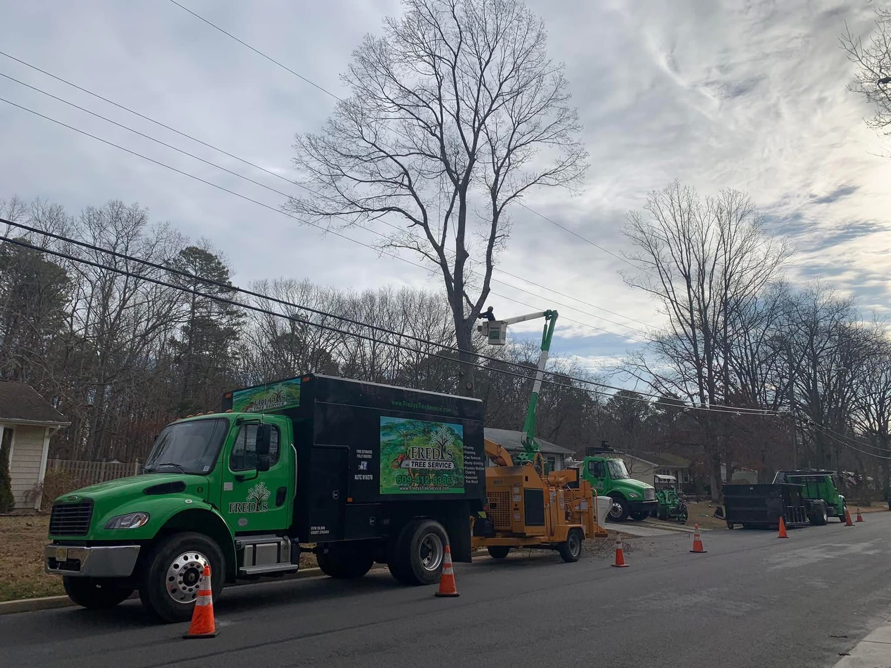 Green utility truck with equipment parked on a street beside cones and trees under cloudy skies