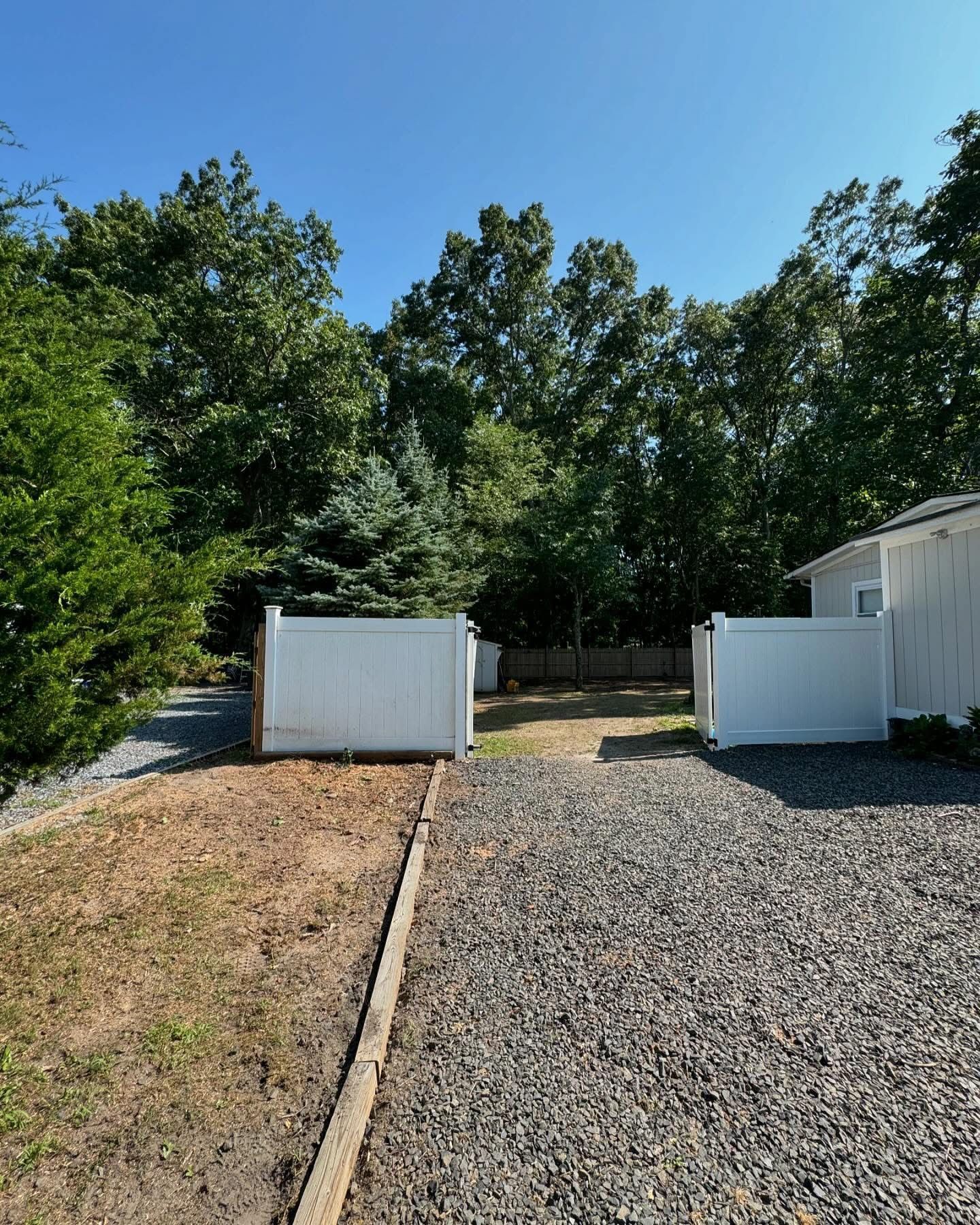 Gravel driveway beside a house, with white fences, trees, and clear blue sky on a sunny day