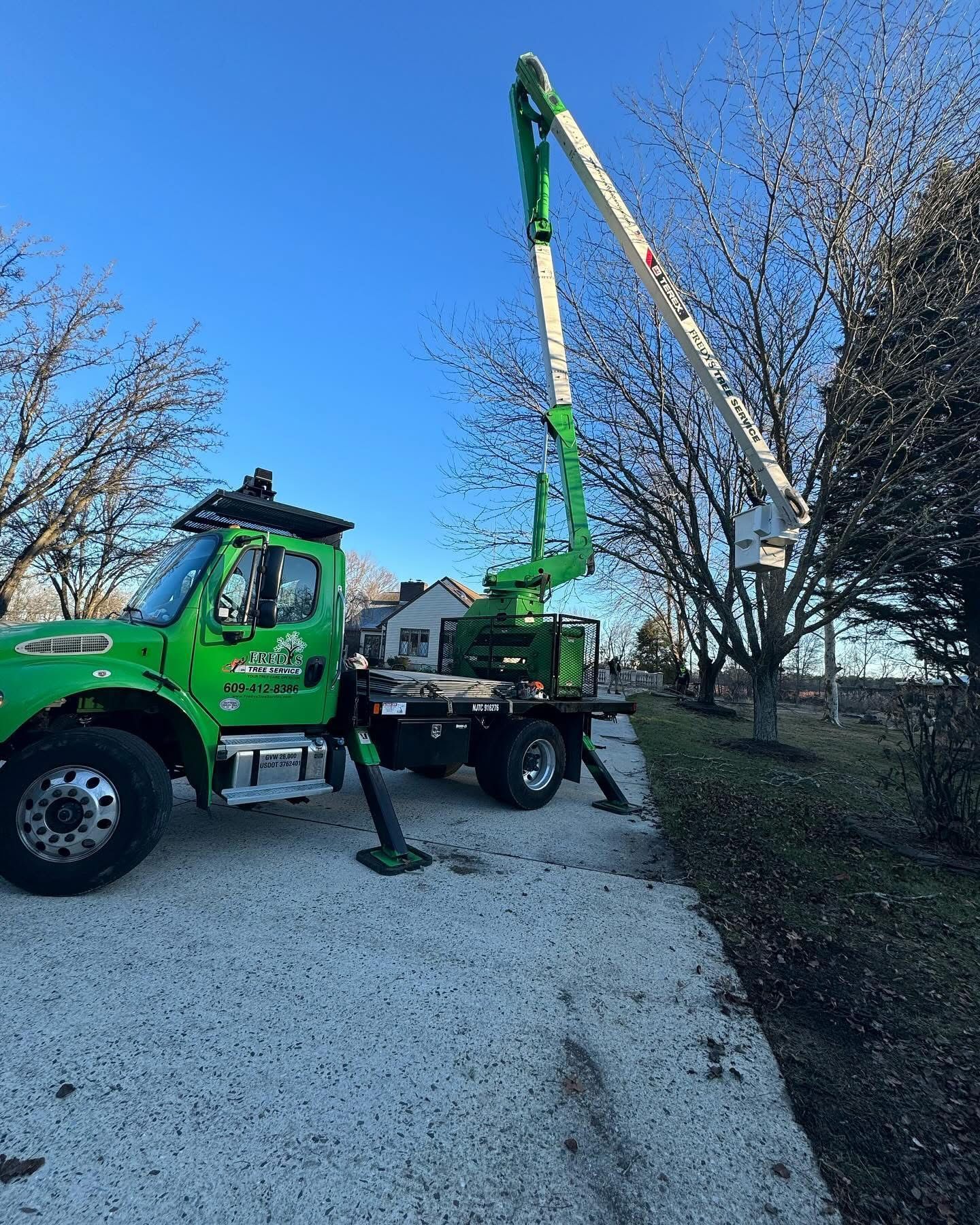 Green utility truck with raised boom lift parked on a gravel driveway beside trees.