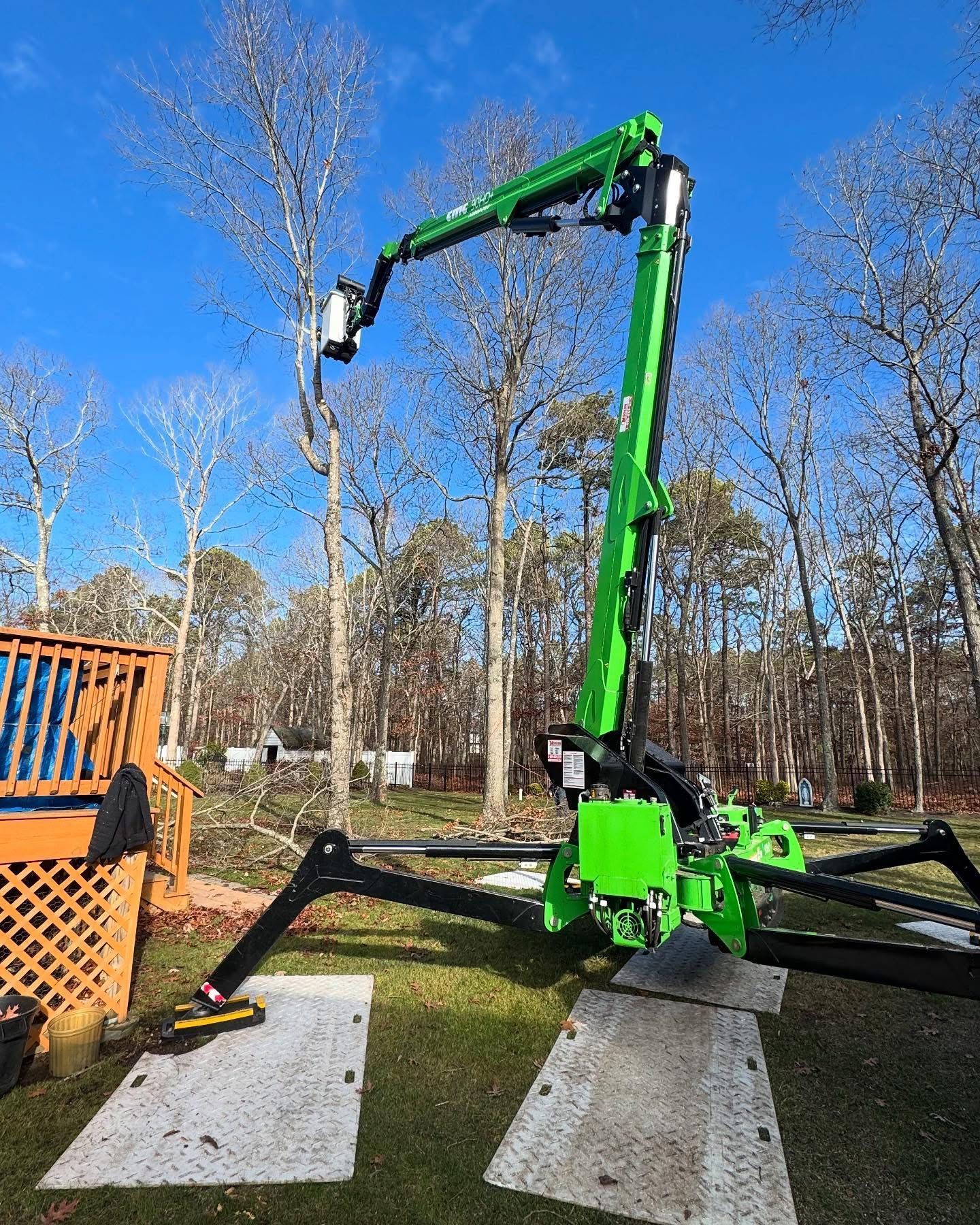 Green excavator arm parked on concrete pads in a grassy yard with trees and blue sky.