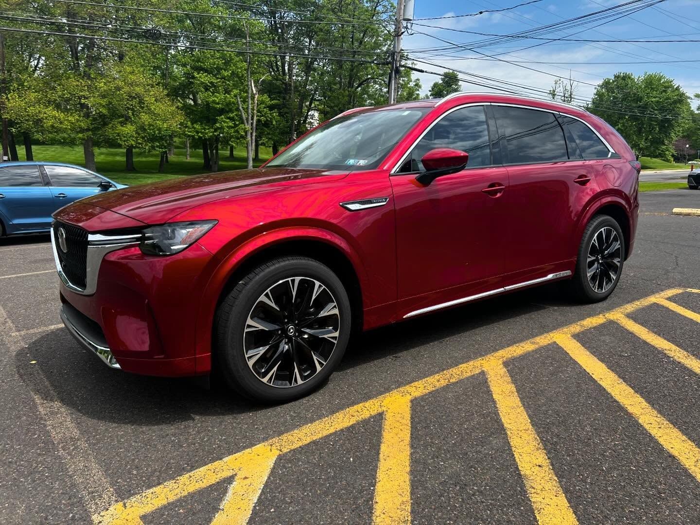 Red Mazda CX-90 SUV parked in a parking space. Black wheels, chrome trim, and tinted windows.