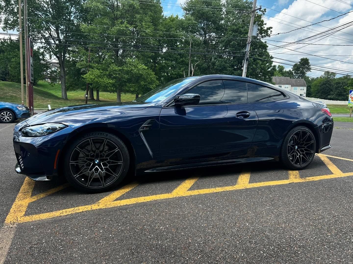 Dark blue BMW coupe parked on yellow-lined pavement. Black rims, tinted windows, trees in the background.