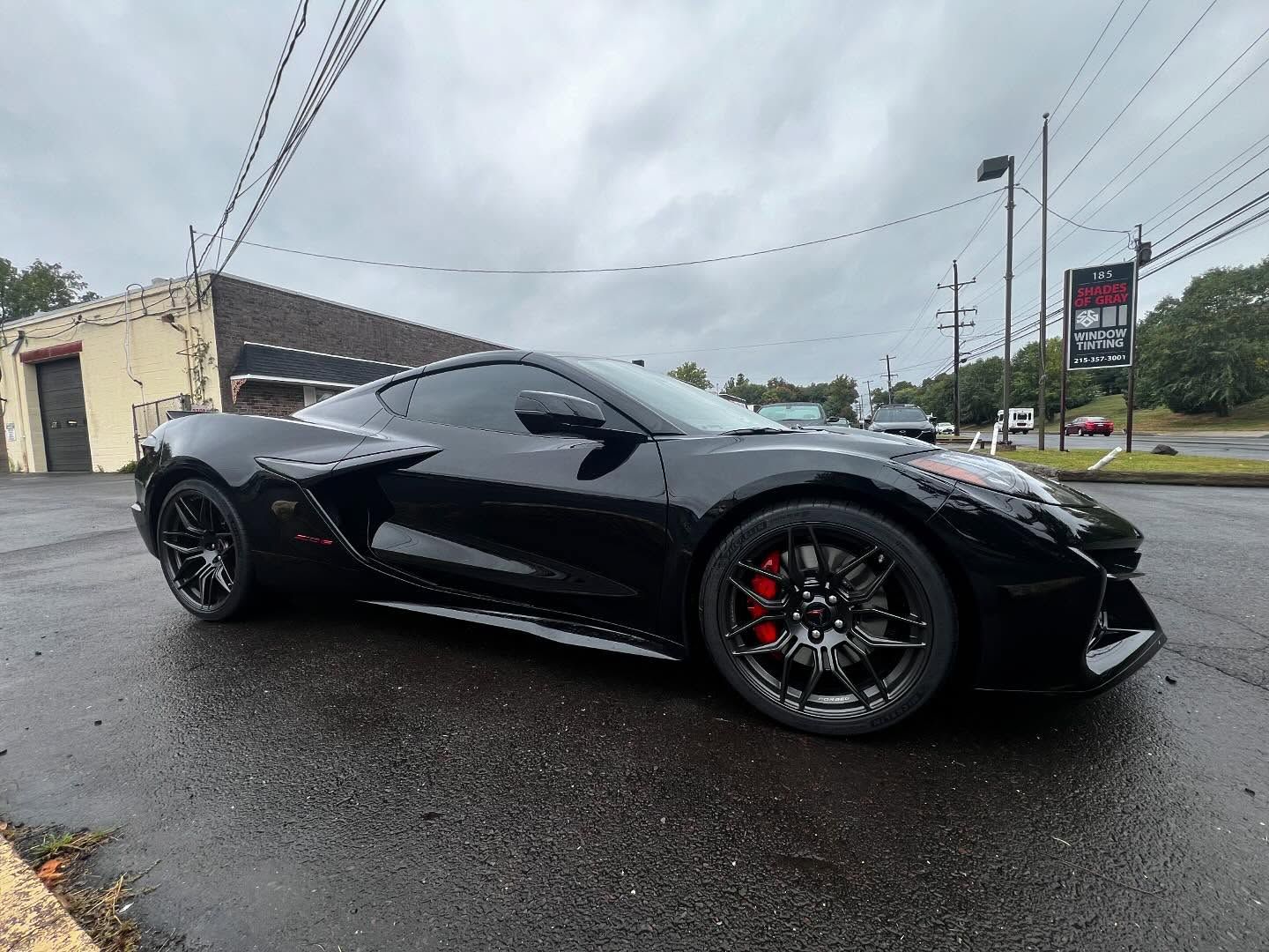 Black Corvette sports car parked on wet asphalt. Red brake calipers visible. Cloudy sky.