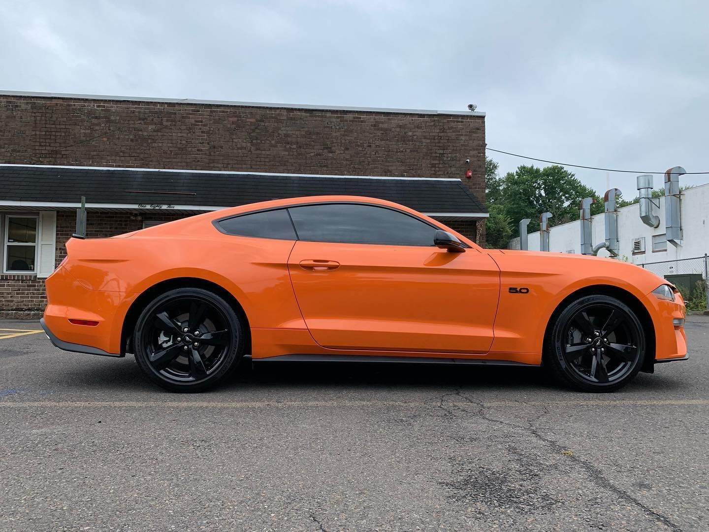 Orange Ford Mustang with black wheels parked in front of a brick building.