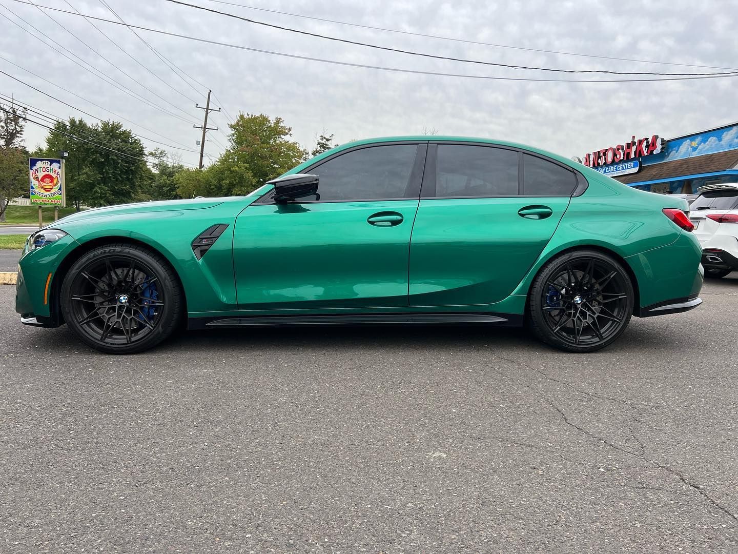 Green BMW M3 sedan with black wheels parked on asphalt in front of a building under a cloudy sky.