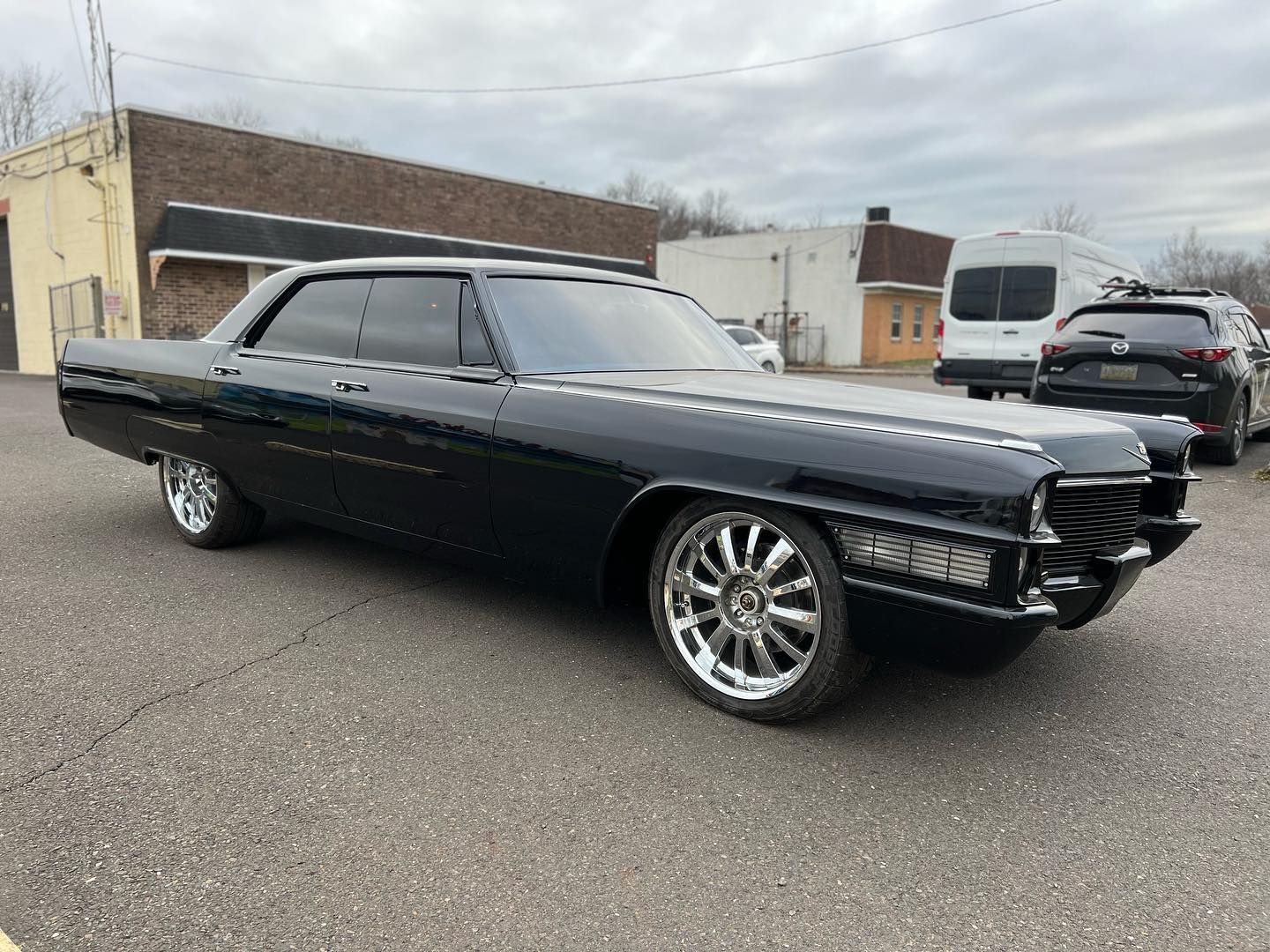 Black Cadillac, chrome wheels, parked on asphalt in front of buildings on a cloudy day.
