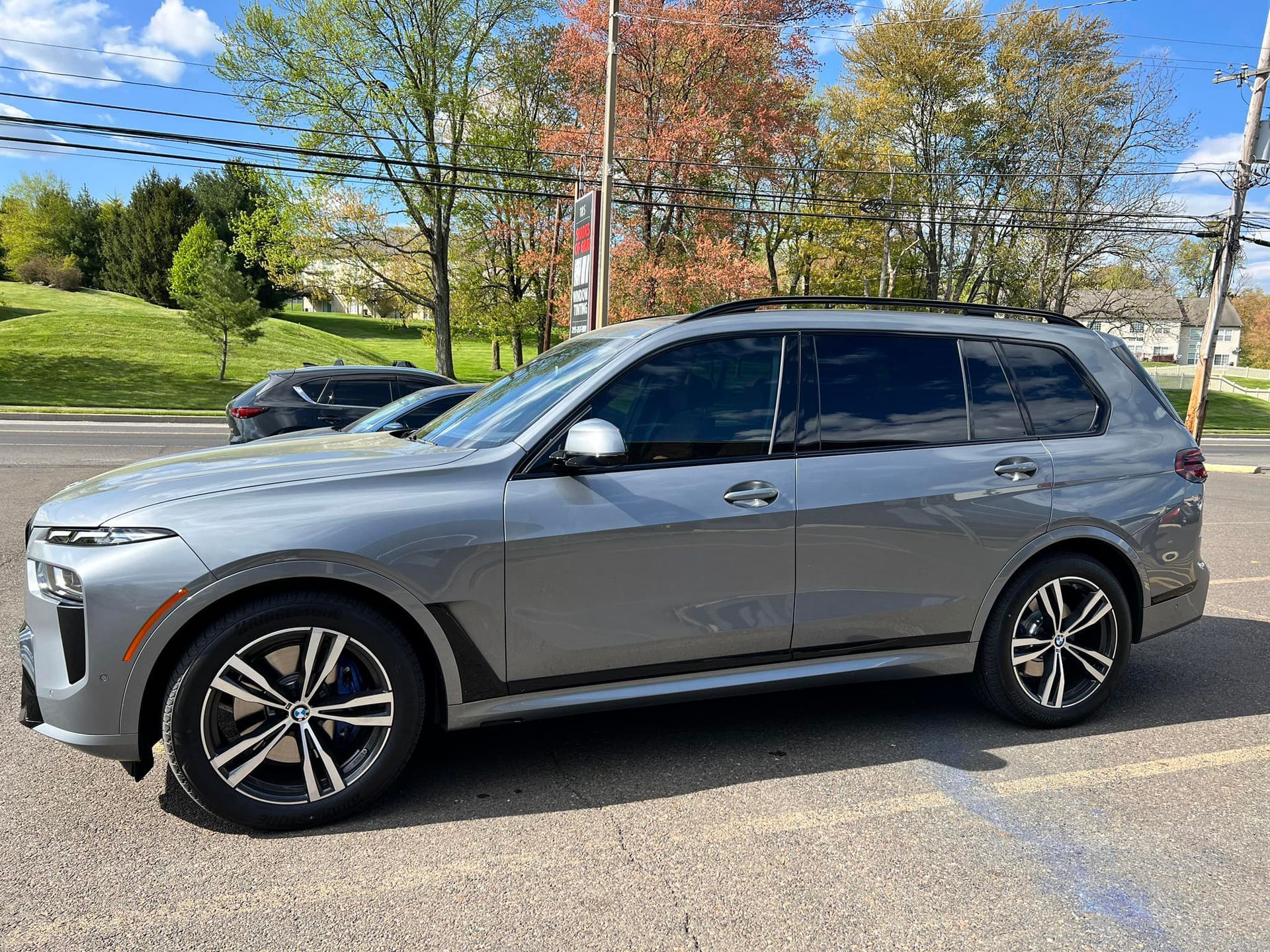 Gray BMW X7 SUV parked on a paved lot with trees and a building in the background.