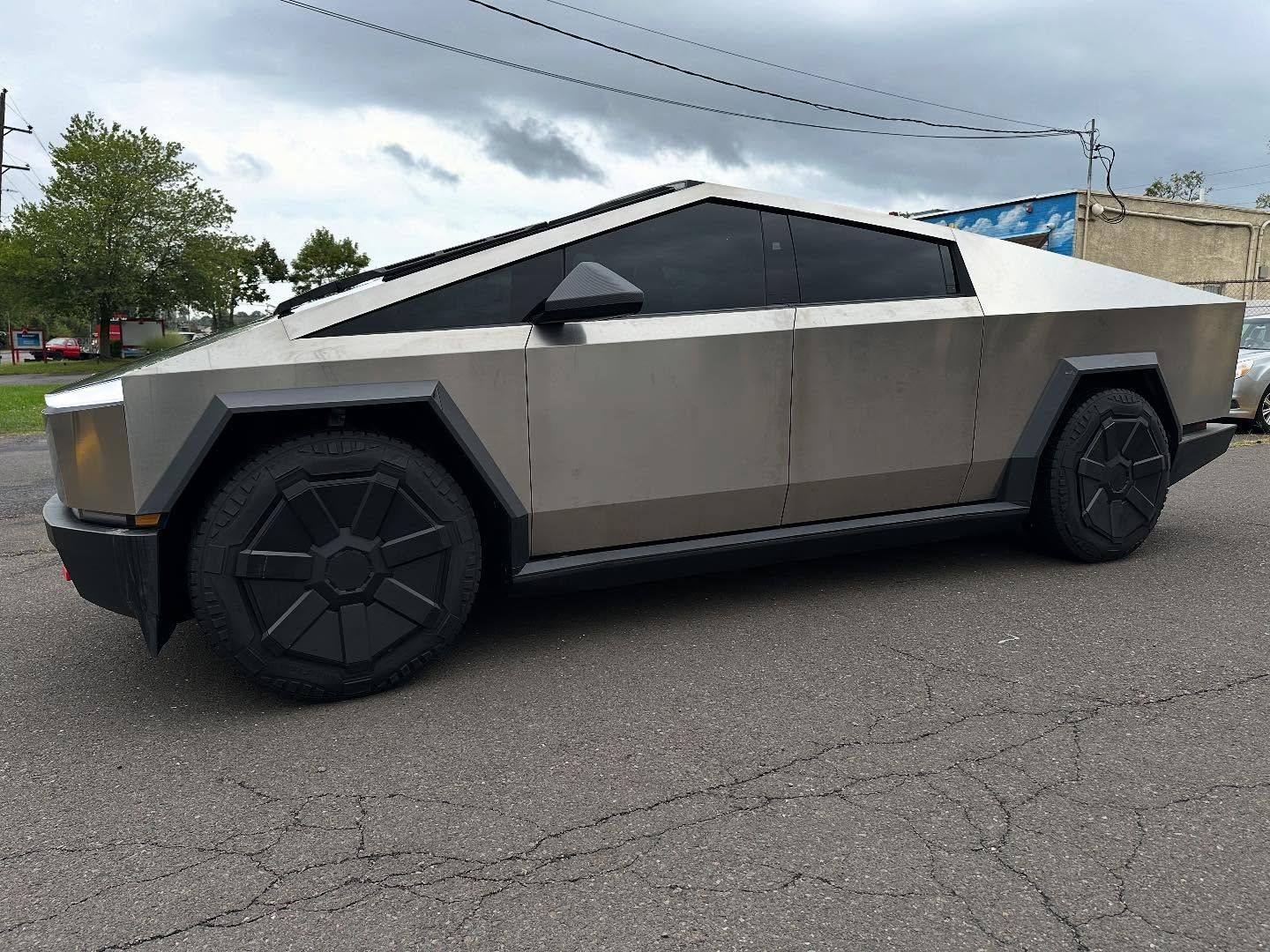 Tesla Cybertruck parked on a paved road, metallic gray exterior, distinctive angular design, black wheels.