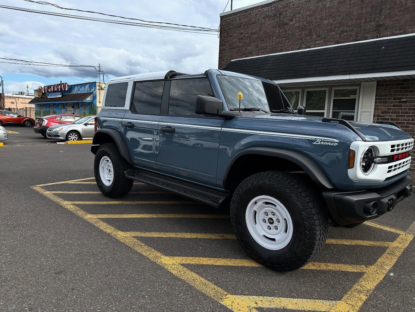 Blue Ford Bronco SUV parked in a yellow-lined space. White wheels, black side steps, and a white roof.