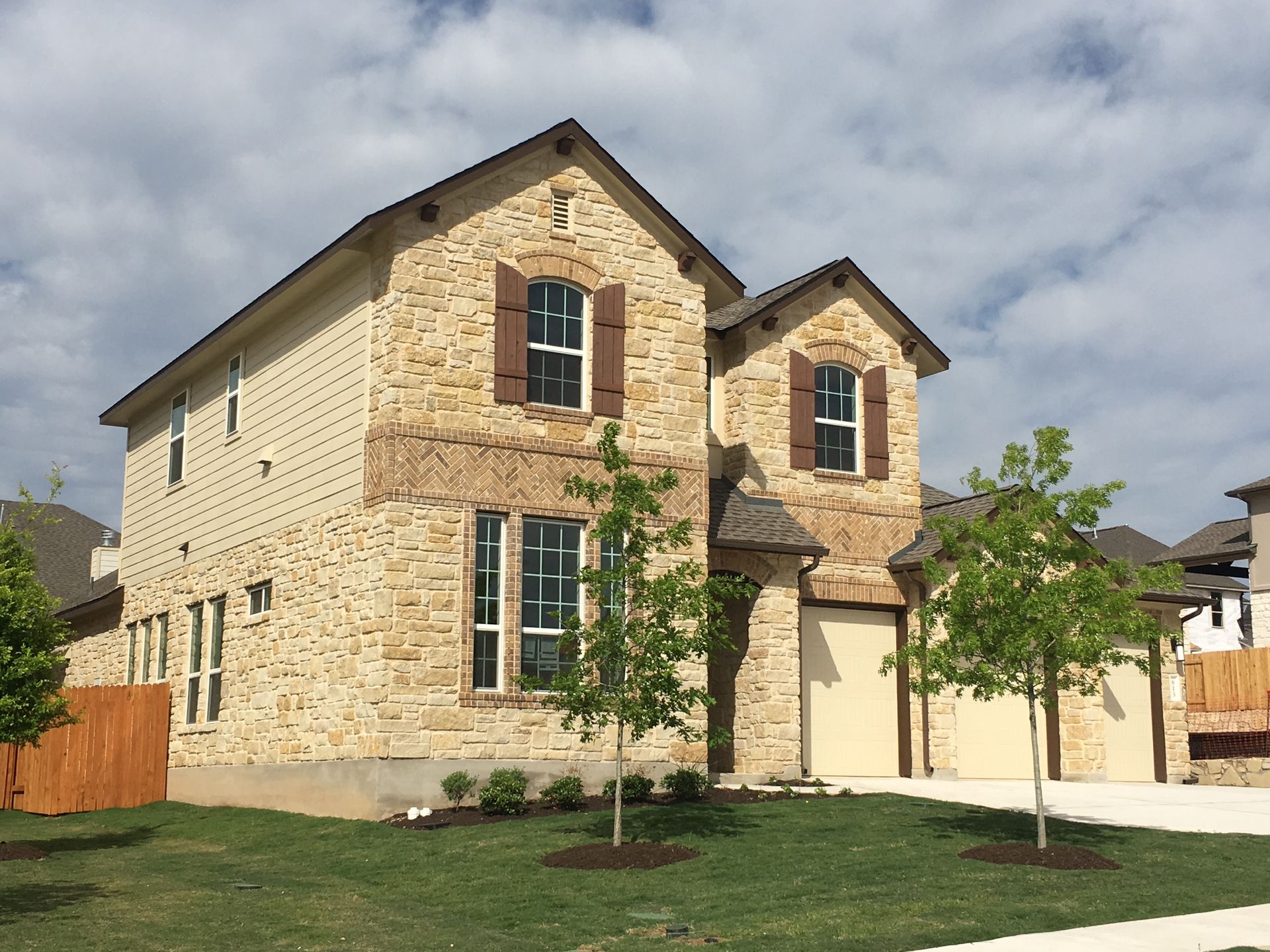 Two-story stone house with brown shutters and a beige garage under a cloudy sky.
