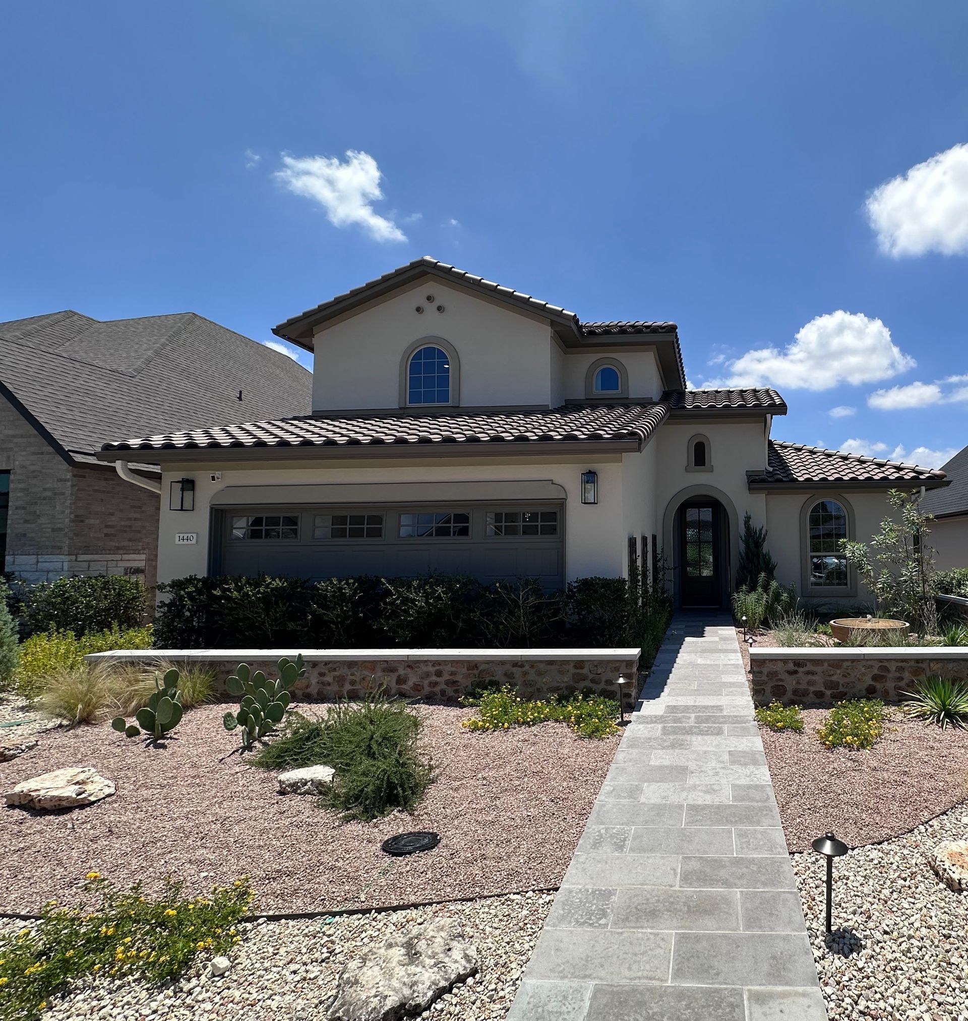Two-story beige stucco house with a tiled roof, stone walkway, and desert landscaping under a blue sky