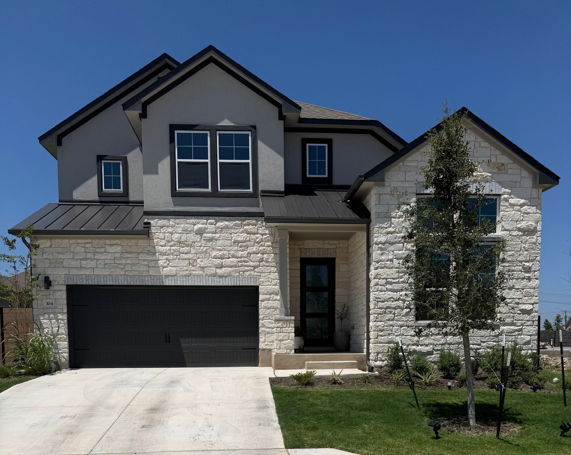 Two-story stone and gray house with a black garage door, front lawn, and driveway under a clear sky