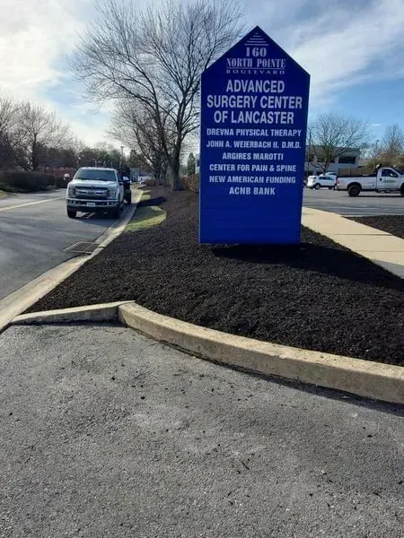 Sign for Advanced Surgery Center of Lancaster; blue, black, and white; beside road; pickup truck in background.