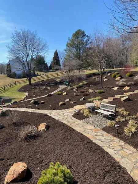 Stone path winds through a landscaped yard with trees, a bench, and a fence under a bright blue sky.