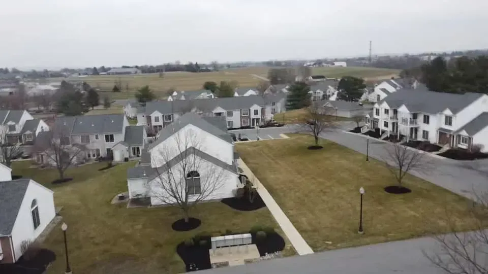 Aerial view of suburban houses with gray roofs on a cloudy day; a field is in the distance.