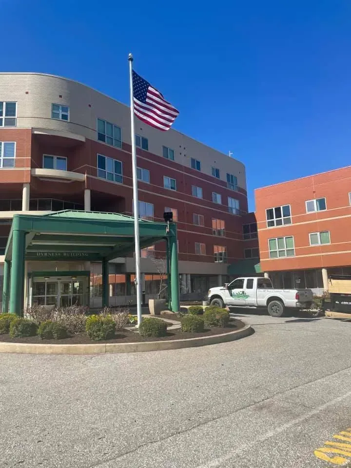 An American flag flies in front of a multistory building with a covered entrance and a white pickup truck parked nearby on a sunny day.