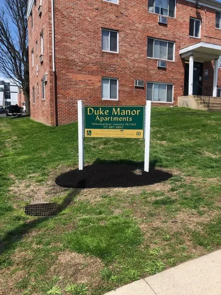 Duke Manor Apartments sign on a grassy area, with a brick apartment building in the background.