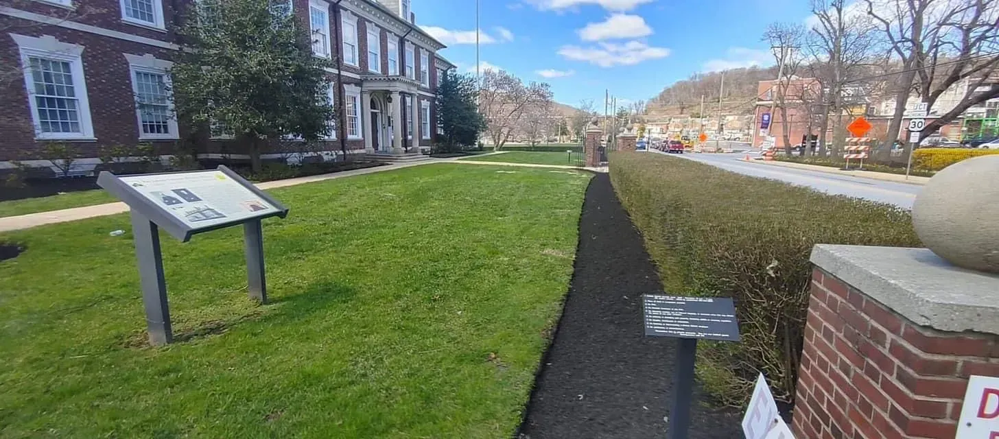 Lush green lawn with a sign. A brick building sits in the background. A path runs along a row of trimmed bushes.