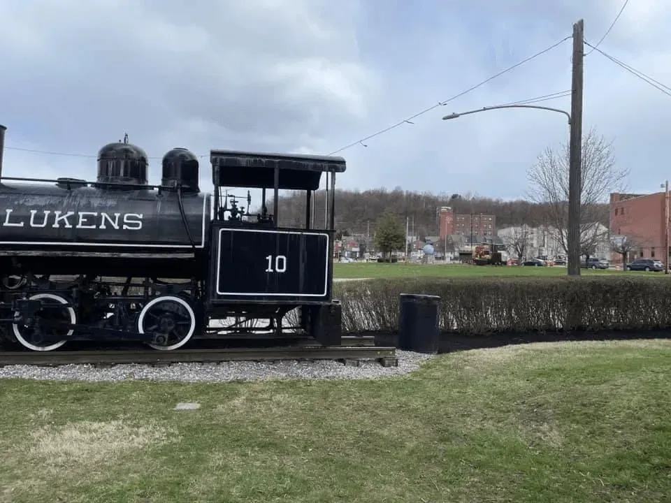 Black Lukens train engine on display in front of a town square with buildings.