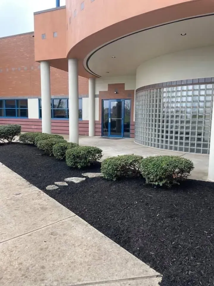 Building entrance with beige pillars, arched roof, and landscaping with bushes and black mulch.