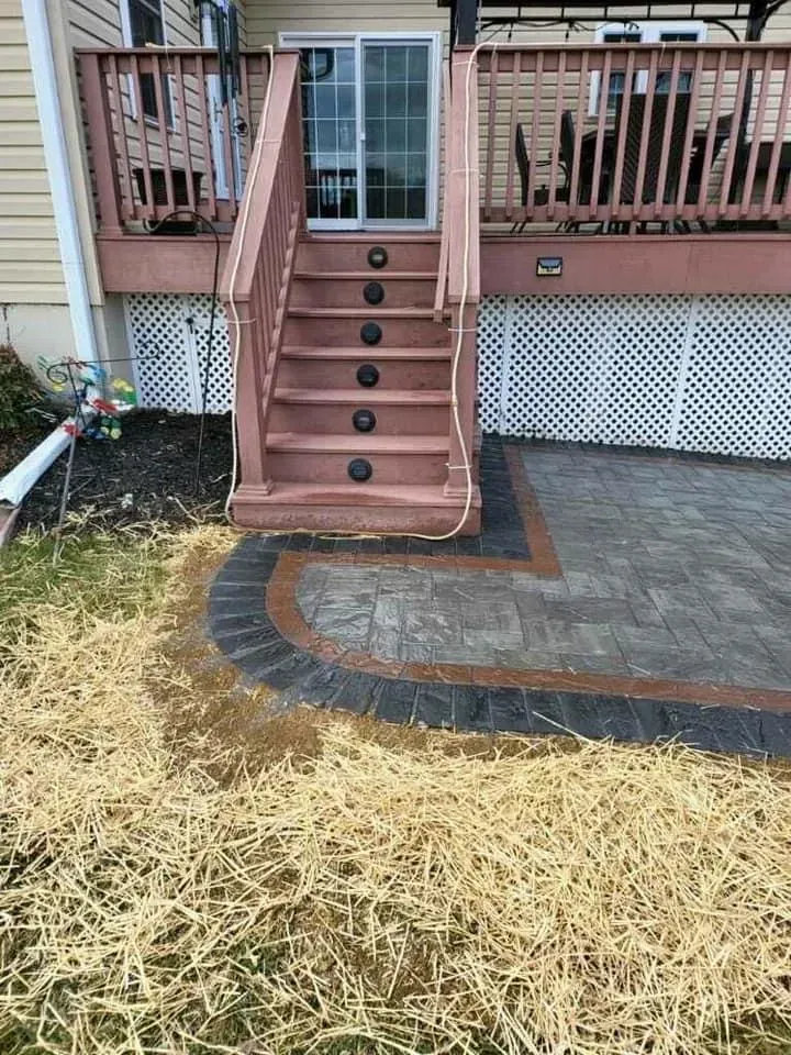 Deck stairs leading to a patio with a brick border, dry grass in the foreground, and a house in the background.