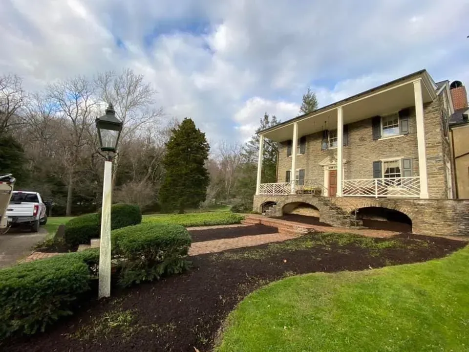 Stone house with porch, walkway, landscaping, and lamp post under cloudy sky.