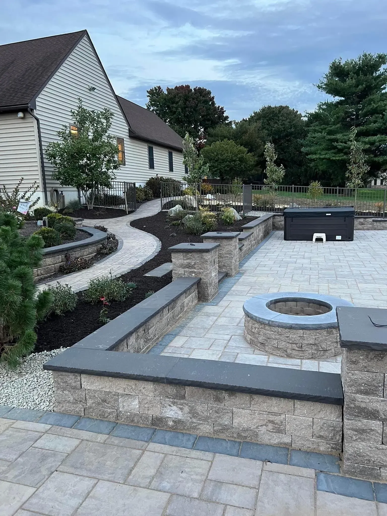 Stone patio with a fire pit, raised walls, and pathway alongside a house with a dark roof.